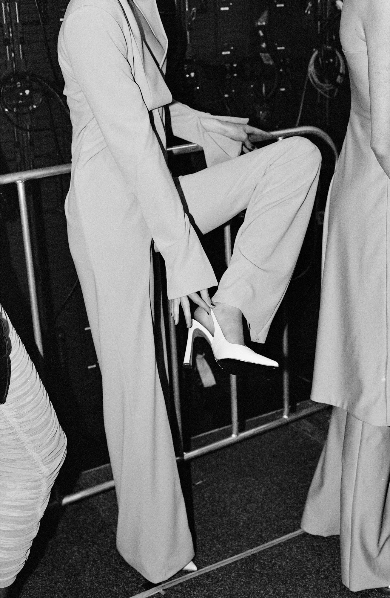 A person wearing a light-colored suit, sitting on a platform with their right leg crossed over their left, wearing high-heeled shoes. The scene appears to be in a backstage or dressing room area, with racks of clothing and equipment visible in the ba