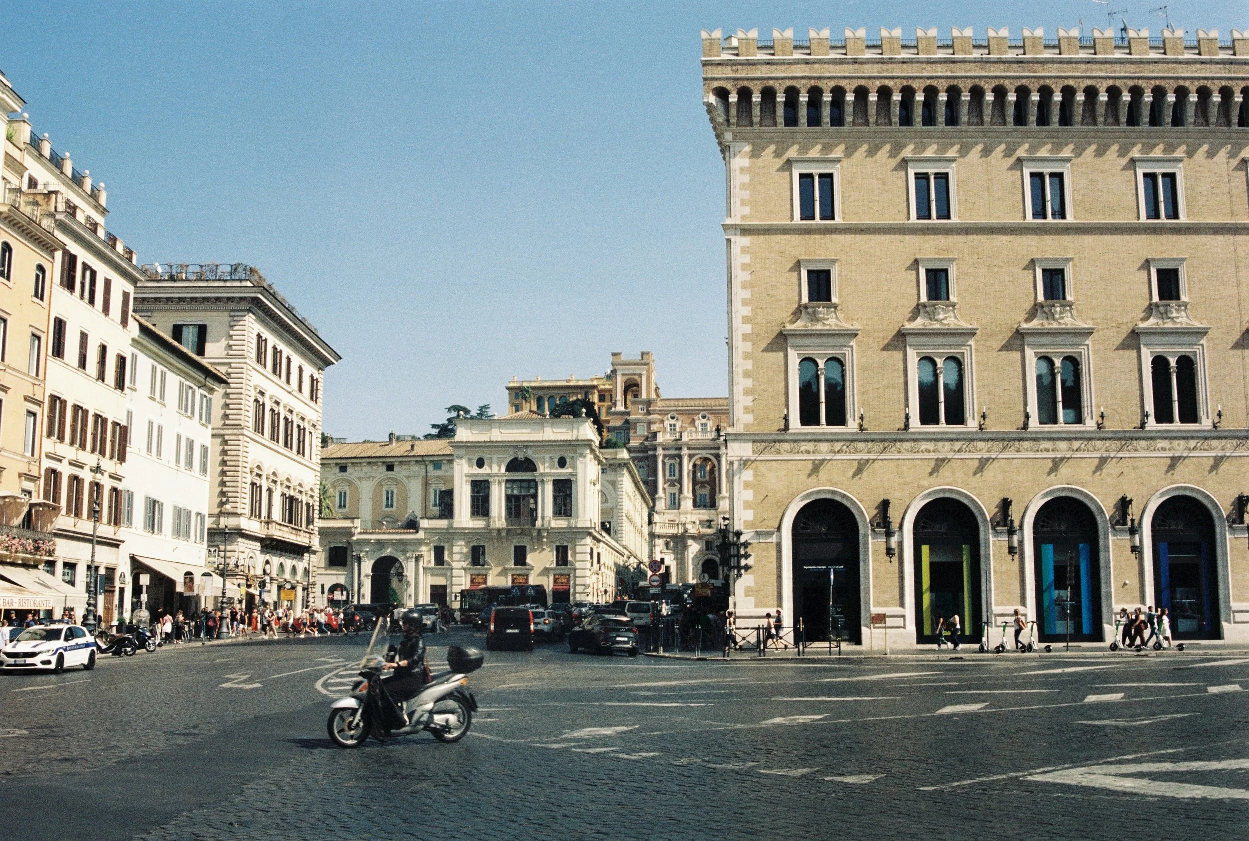 A busy city square with cars, motorcycles, and pedestrians. Elegant historic buildings with arched windows and ornate details line the square, and a large beige building with decorative balconies dominates the scene.