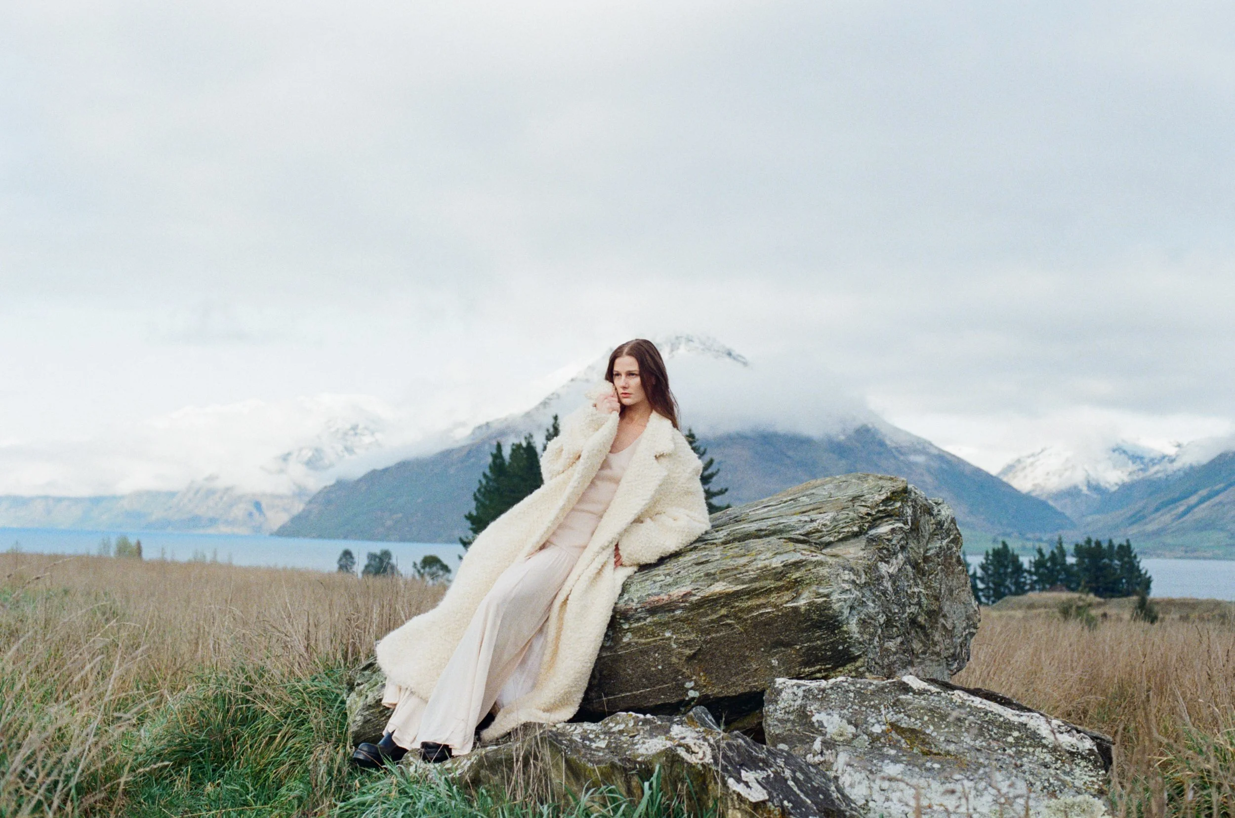 A woman sitting on a large rock outdoors with mountains, a lake, and cloudy sky in the background. She is wearing a cream-colored wool coat and light-colored pants.