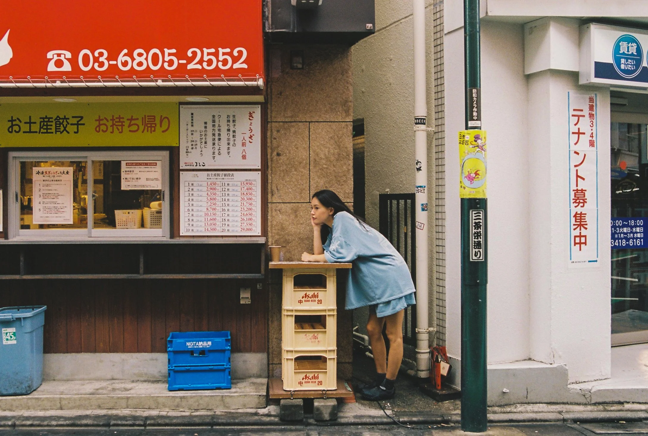 A young girl wearing a blue dress and black shoes is leaning on a wooden table outside a shop, looking to her right. The shop has signs in Japanese, and there are plastic crates and bins below the window, with one marked 'Asahi' beer.