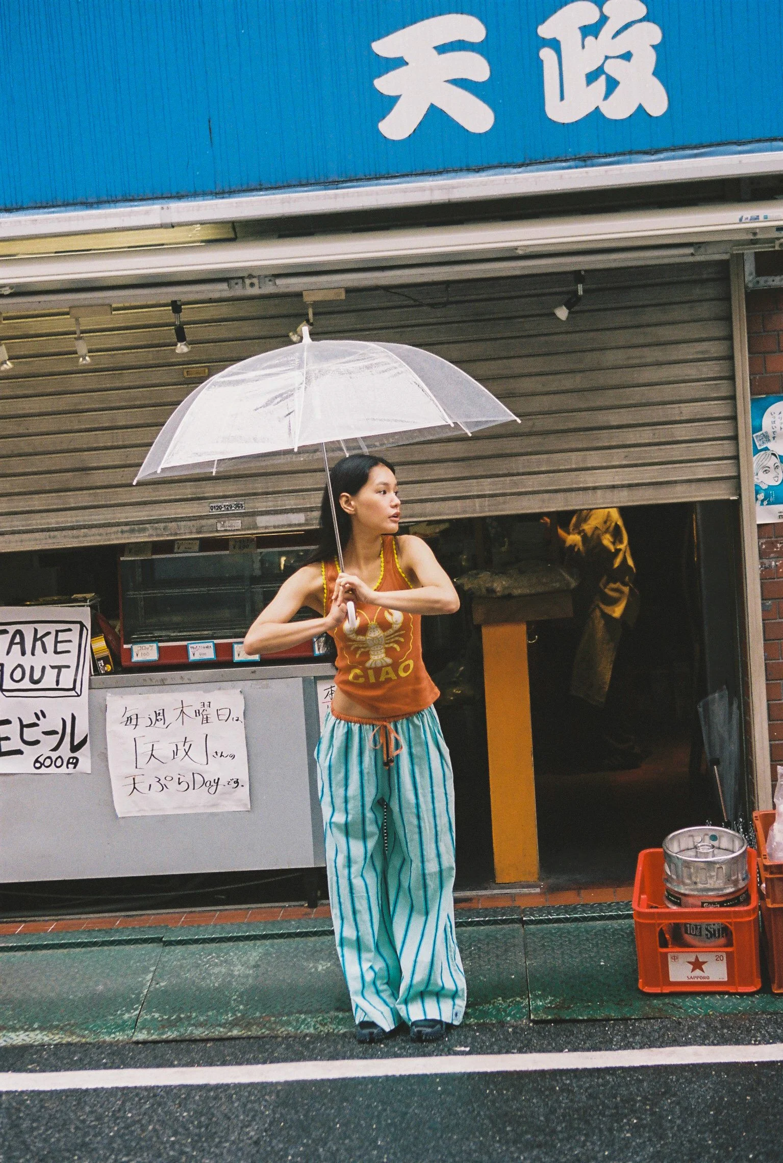 A woman with short dark hair holding a clear umbrella, standing in front of a closed shop with Japanese signs. She is wearing a sleeveless orange top with a dragon design and striped turquoise and white wide-leg pants. There are handwritten signs on 