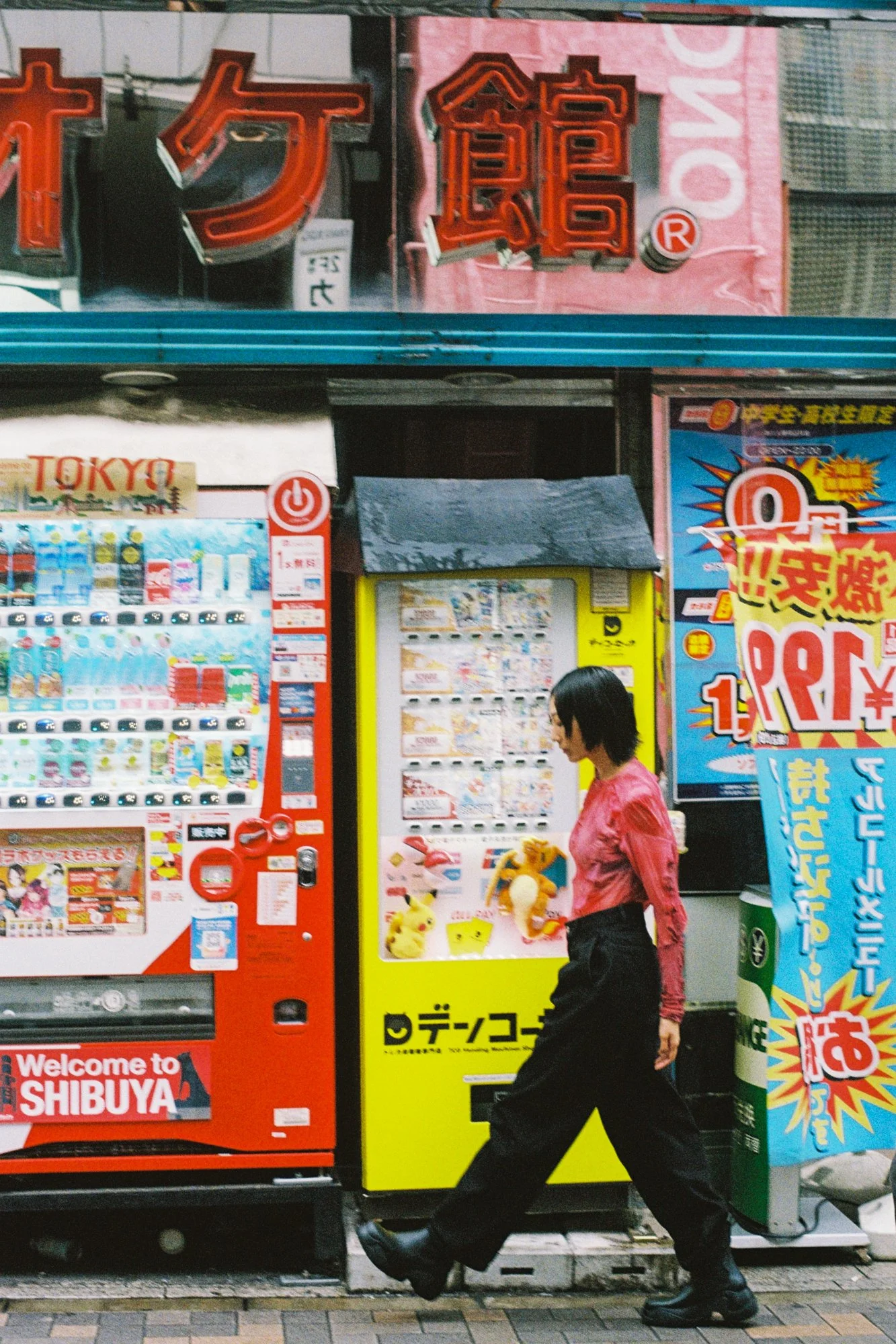 A woman in a pink jacket and black pants walking past a vending machine and a yellow and black sign with Japanese characters outside in an urban area. The vending machine displays Pokémon plush toys.