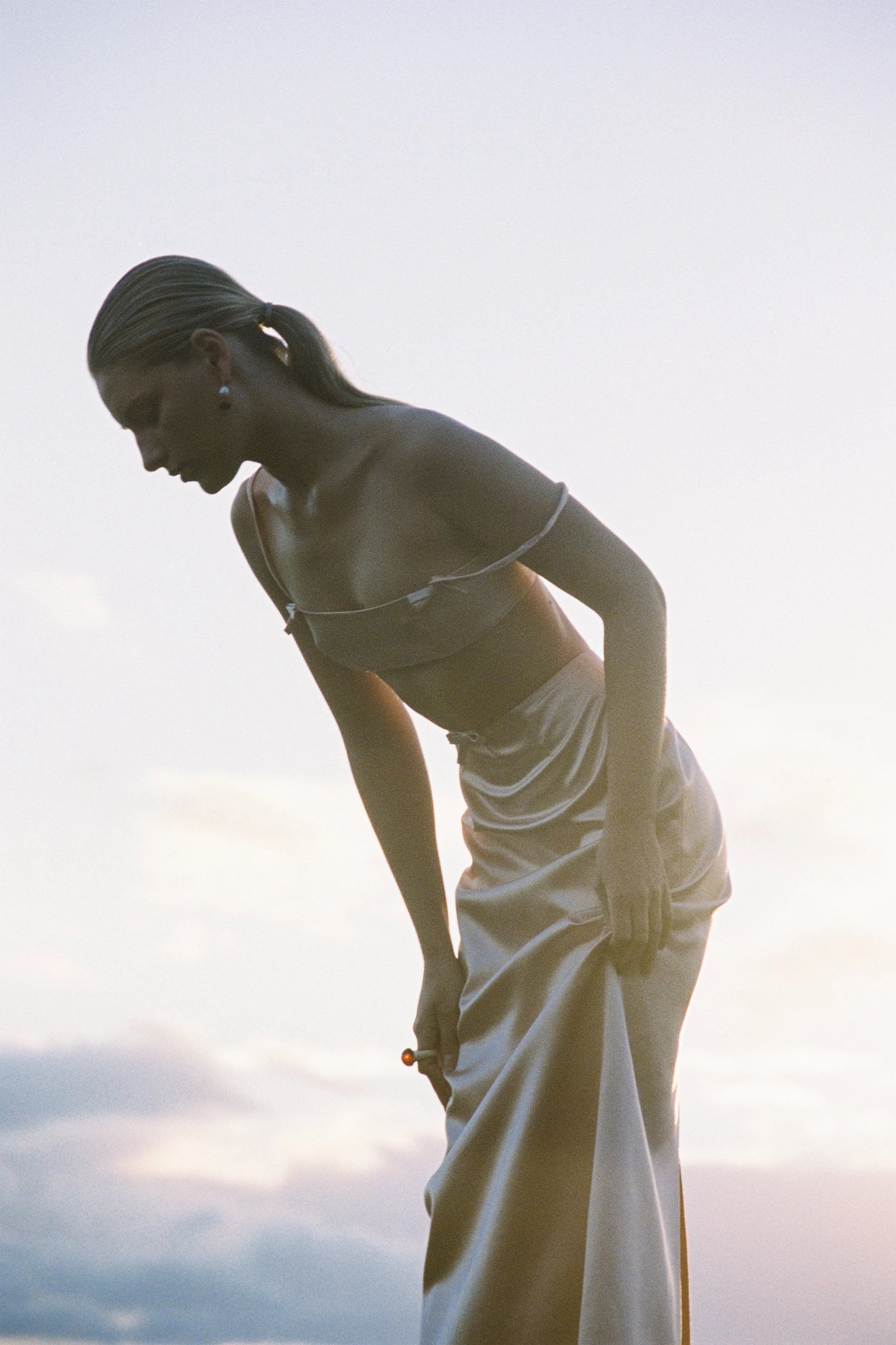 A person wearing a tank top and sweatpants, with a ponytail, is leaning forward against the sky at sunset or sunrise.