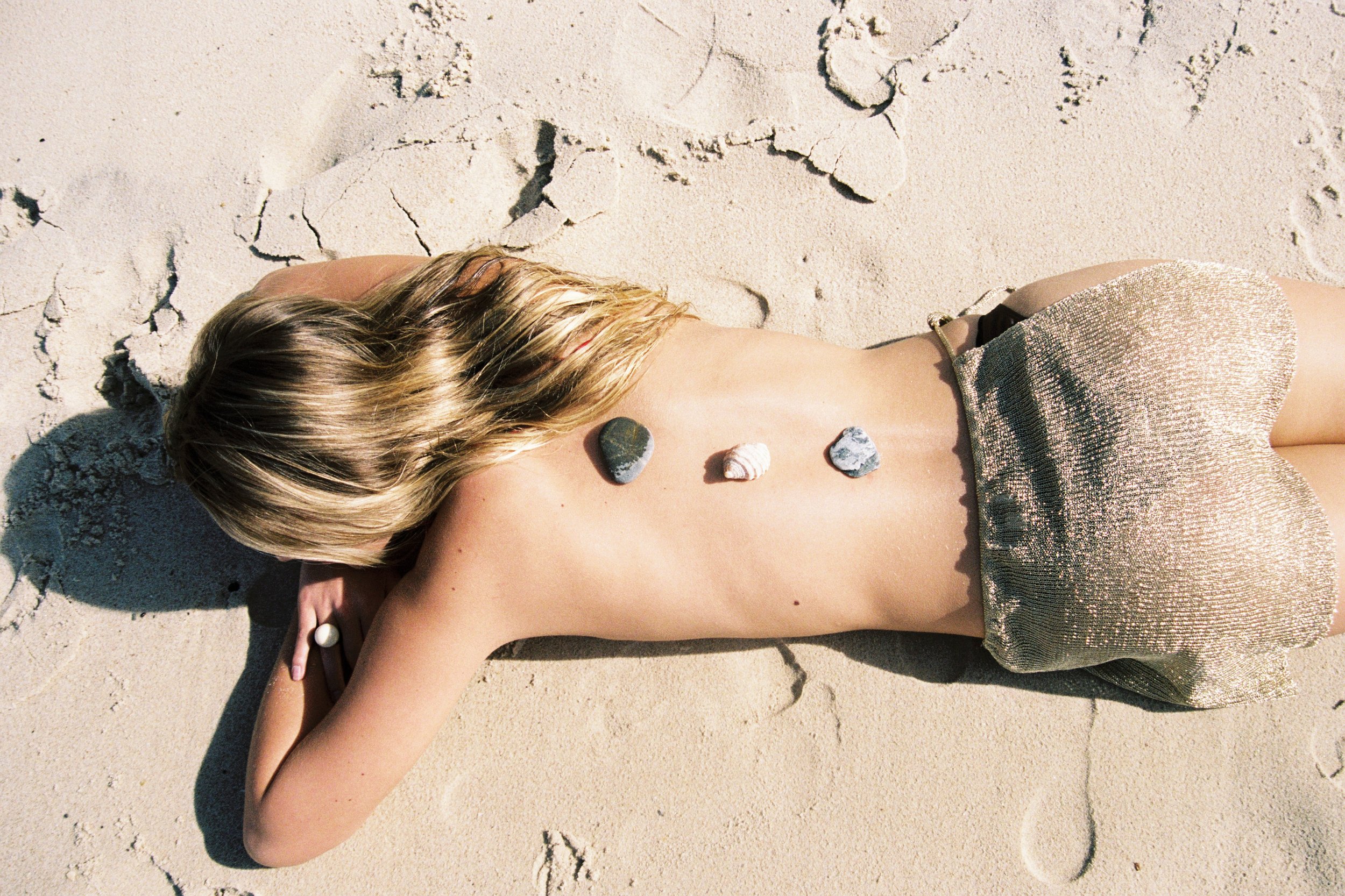 A woman is lying face down on sandy ground, with her blonde hair spread out and wearing a shiny, metallic dress. There are three seashells placed on her back.