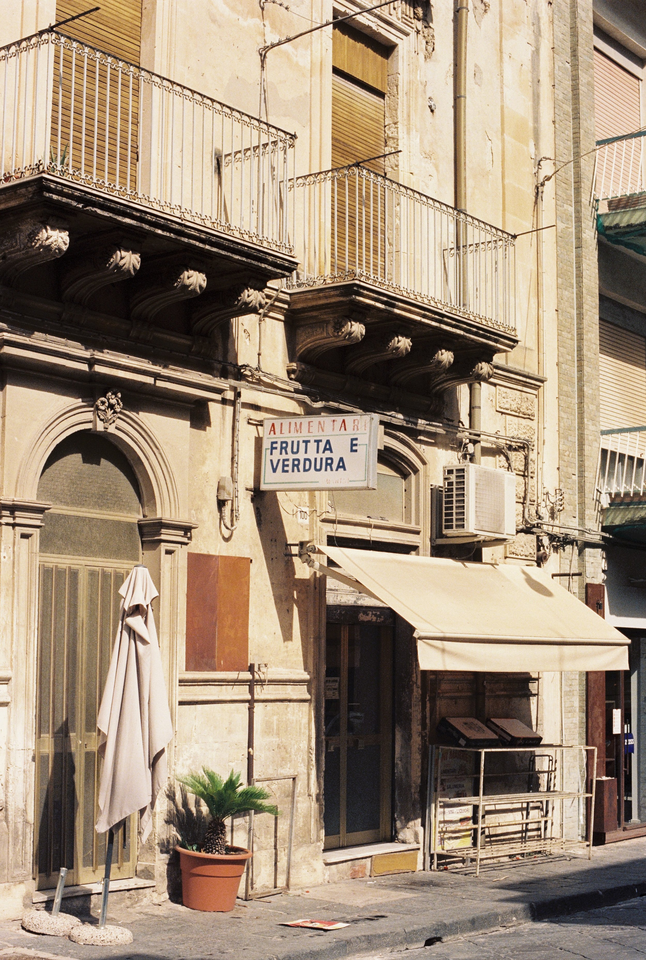 Sunlit city street scene featuring a building facade with balconies, shuttered windows, a storefront with a sign reading 'Frutta e Verdura,' potted plants, a folded umbrella, and building details like ornate cornices and air conditioning units.