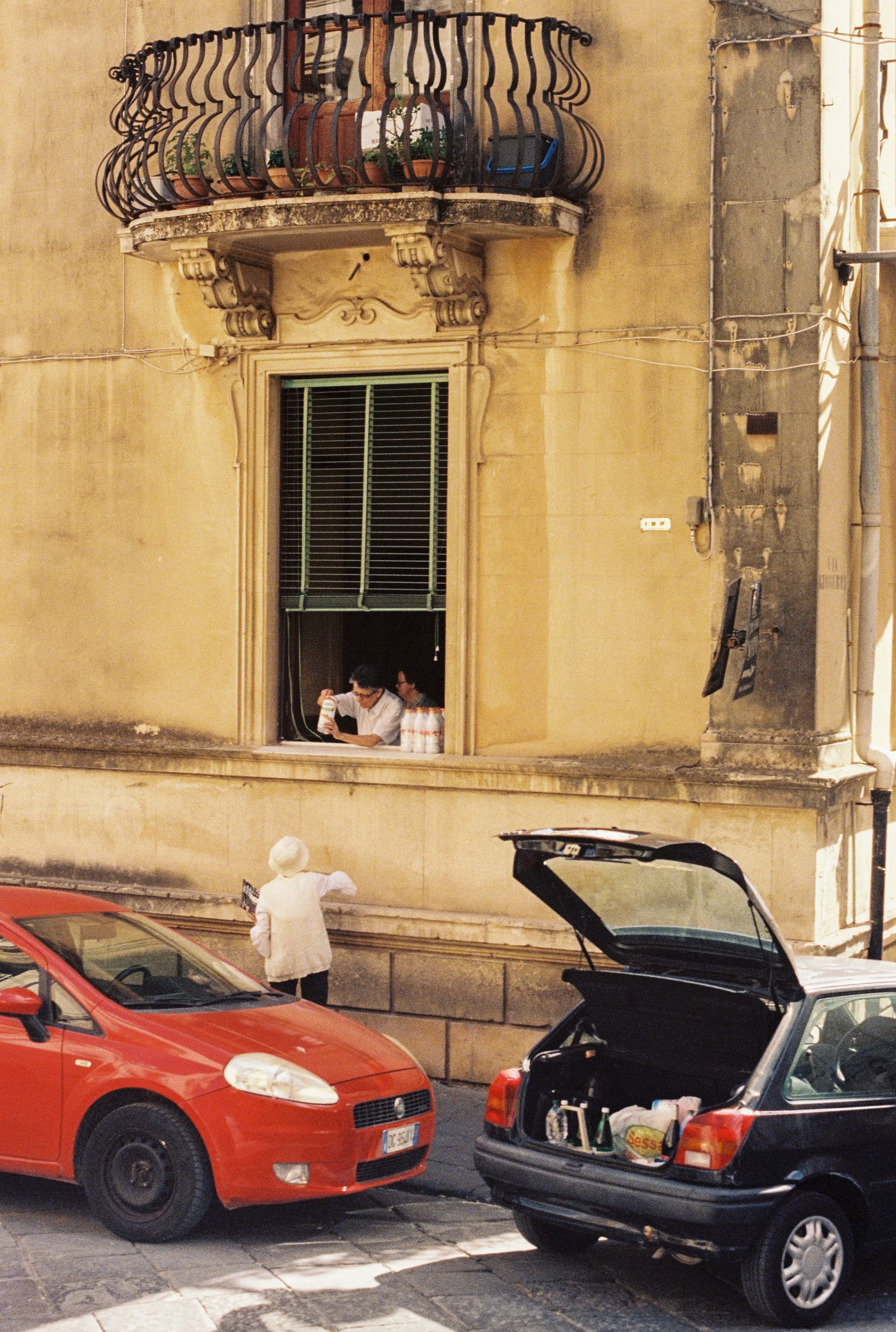 A street scene in Italy with parked cars, including a red car and a black car with an open trunk. An old lady is lifting a bag of groceries. The background consists of an old yellow building with a small balcony and a window with shutters.