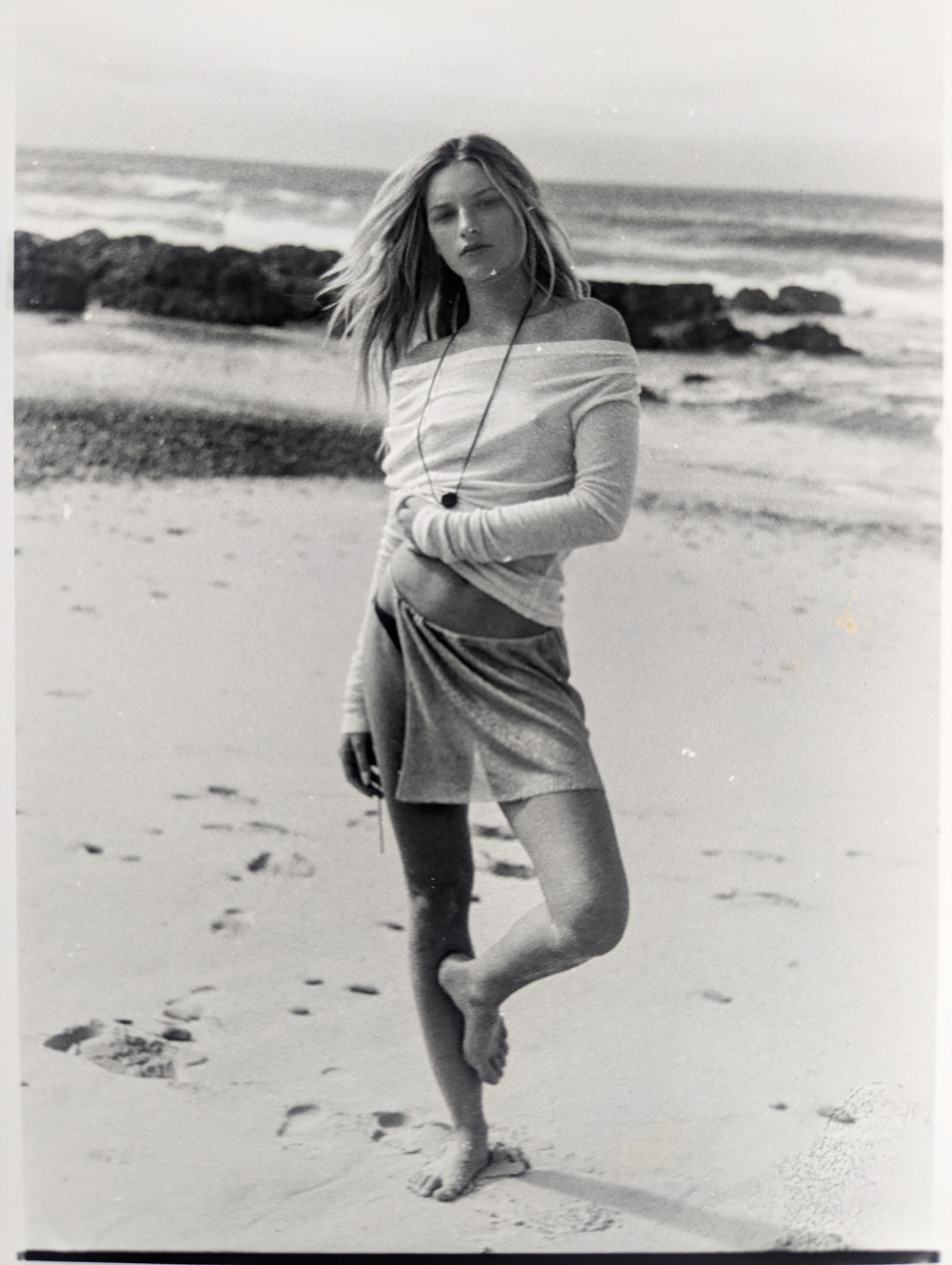 A black-and-white photo of a woman standing on a beach near the water, with rocks in the background. She has shoulder-length hair and is wearing a long-sleeved shirt and a skirt, with a necklace. She is barefoot, looking at the camera with a serious 