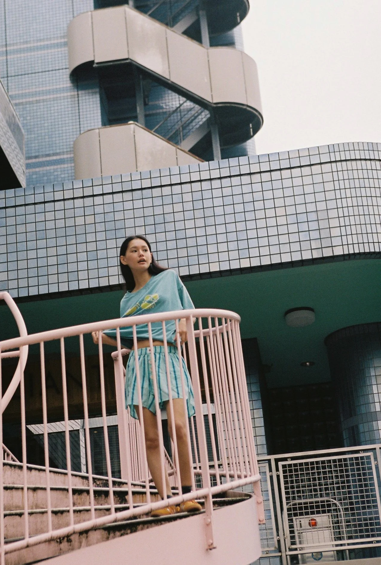 A young woman with long dark hair standing on a pink balcony railing outside a building with a tiled exterior. She is wearing a light blue t-shirt and striped shorts, looking to the side.