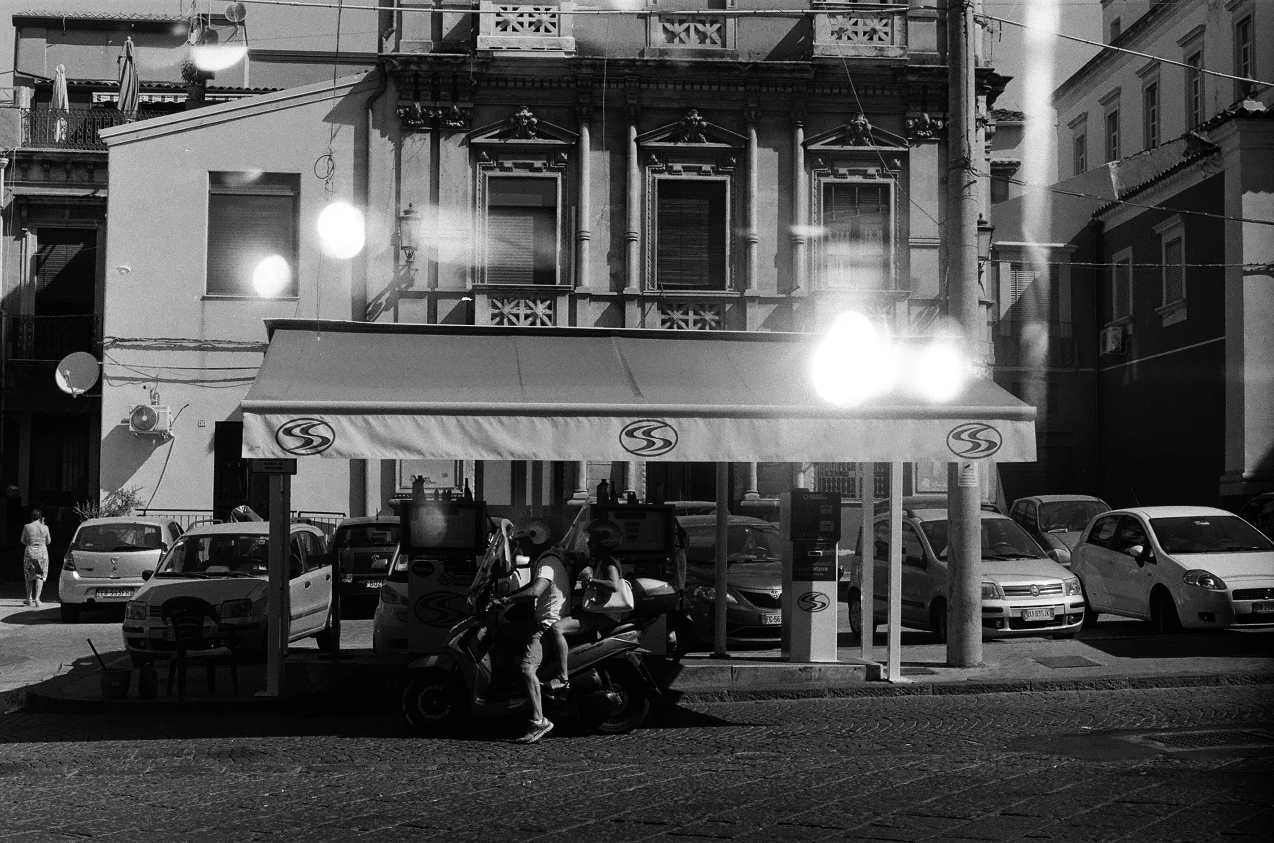 Black and white photo of a city street with cars parked along the curb, a fast food kiosk with a canopy, a pedestrian, and older buildings in the background.