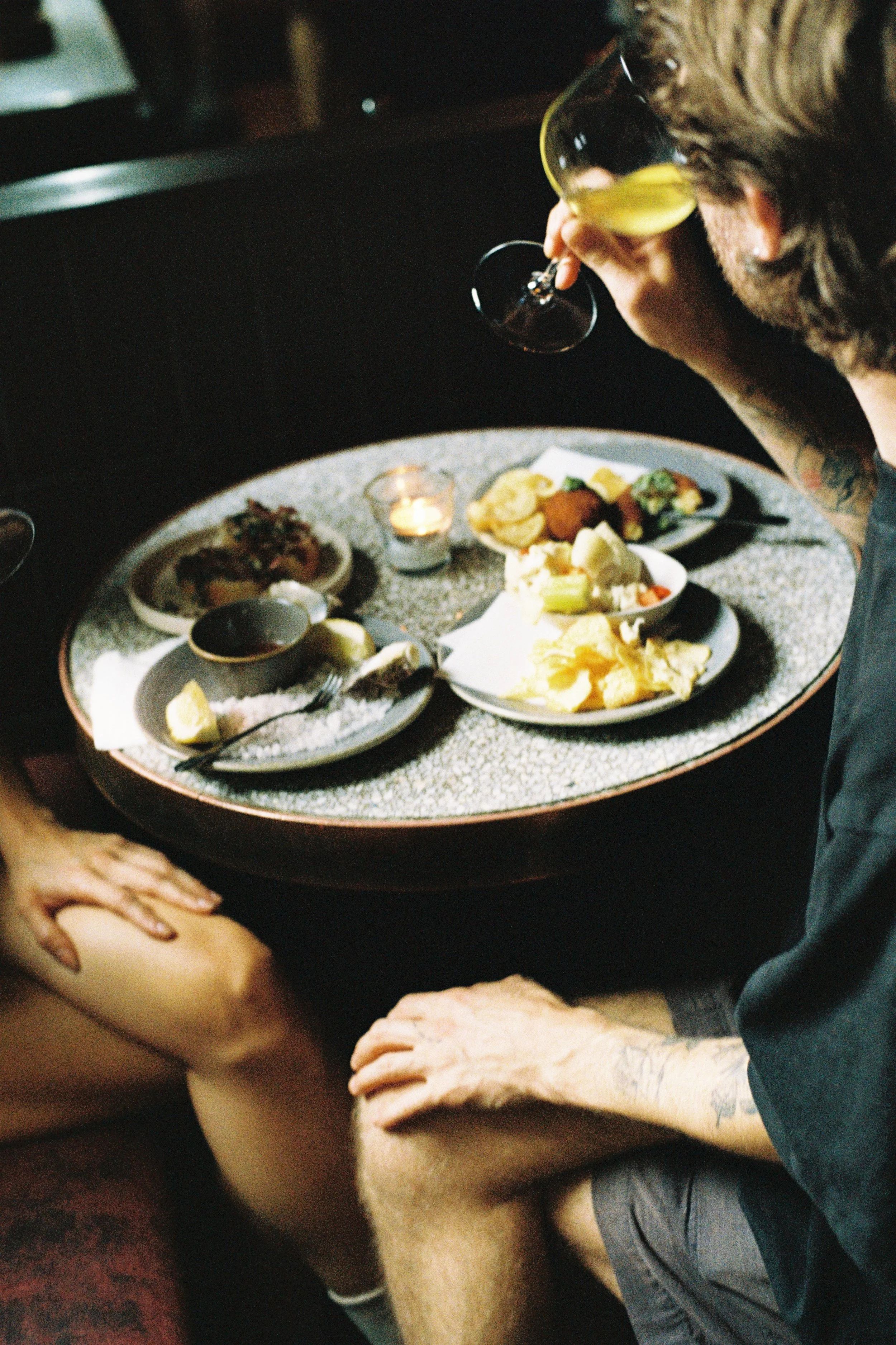 A person with tattoos on their arms drinking from a wine glass at a dining table with various dishes and a lit candle.