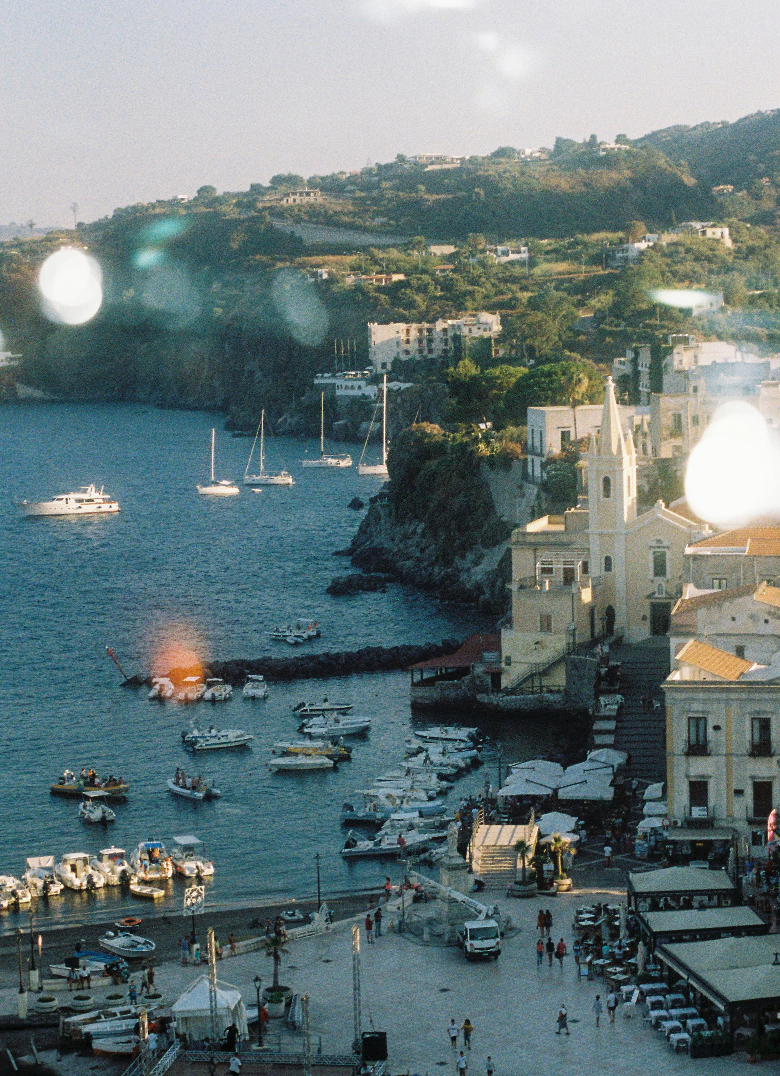 A picturesque harbor scene with boats docked in the water, a church with a steeple, and buildings along the shoreline. Hills with houses and greenery rise in the background, and people are walking along the waterfront promenade.