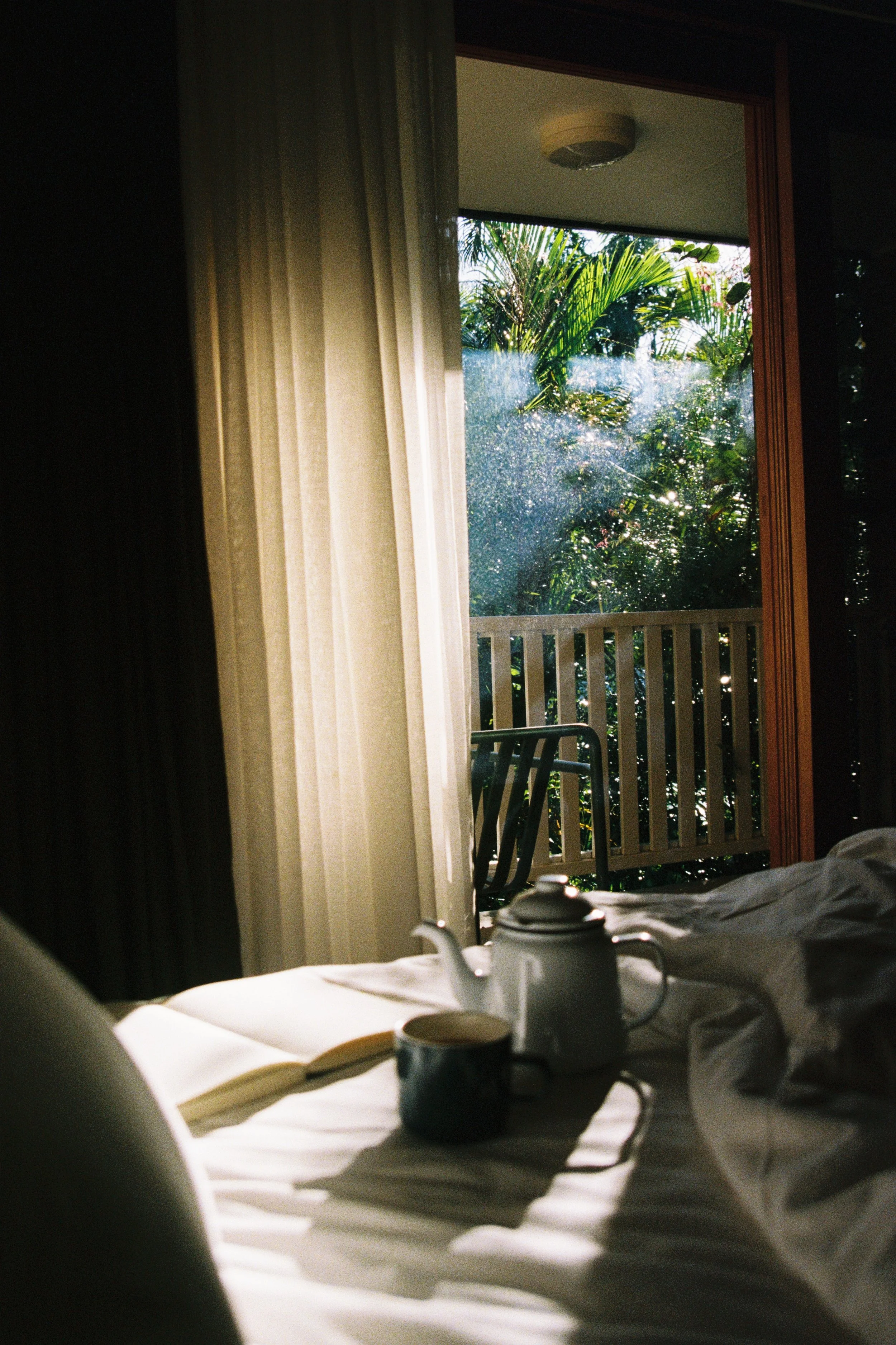 A view from a bed in a hotel room showing a balcony with outdoor chairs and lush green tropical plants outside.