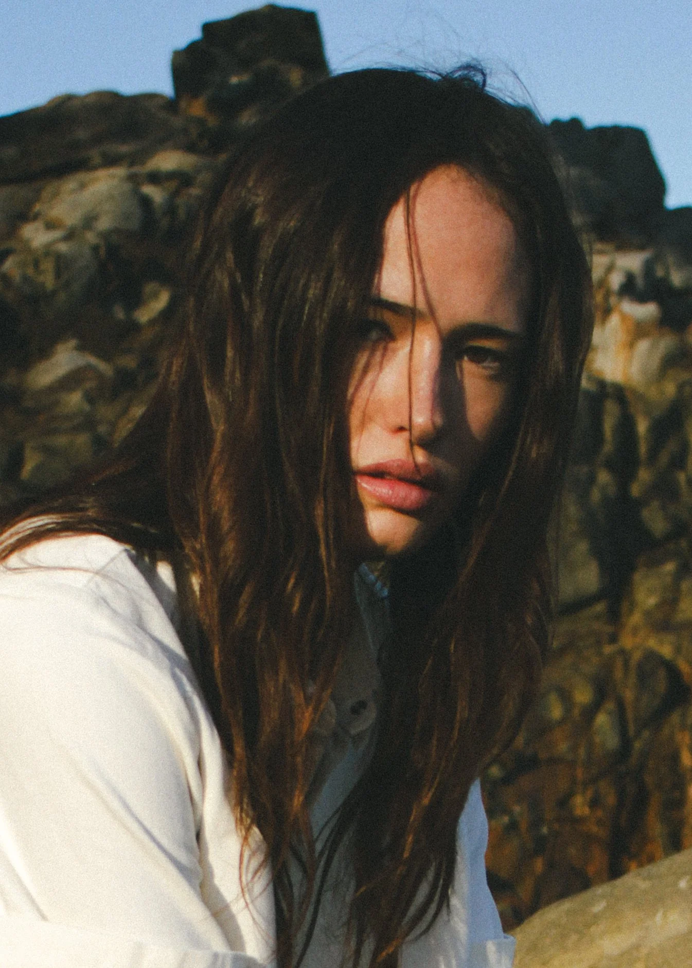 Close-up of a woman with long brunette wavy hair, looking down and slightly to the side, with rocks in the background and a clear sky.