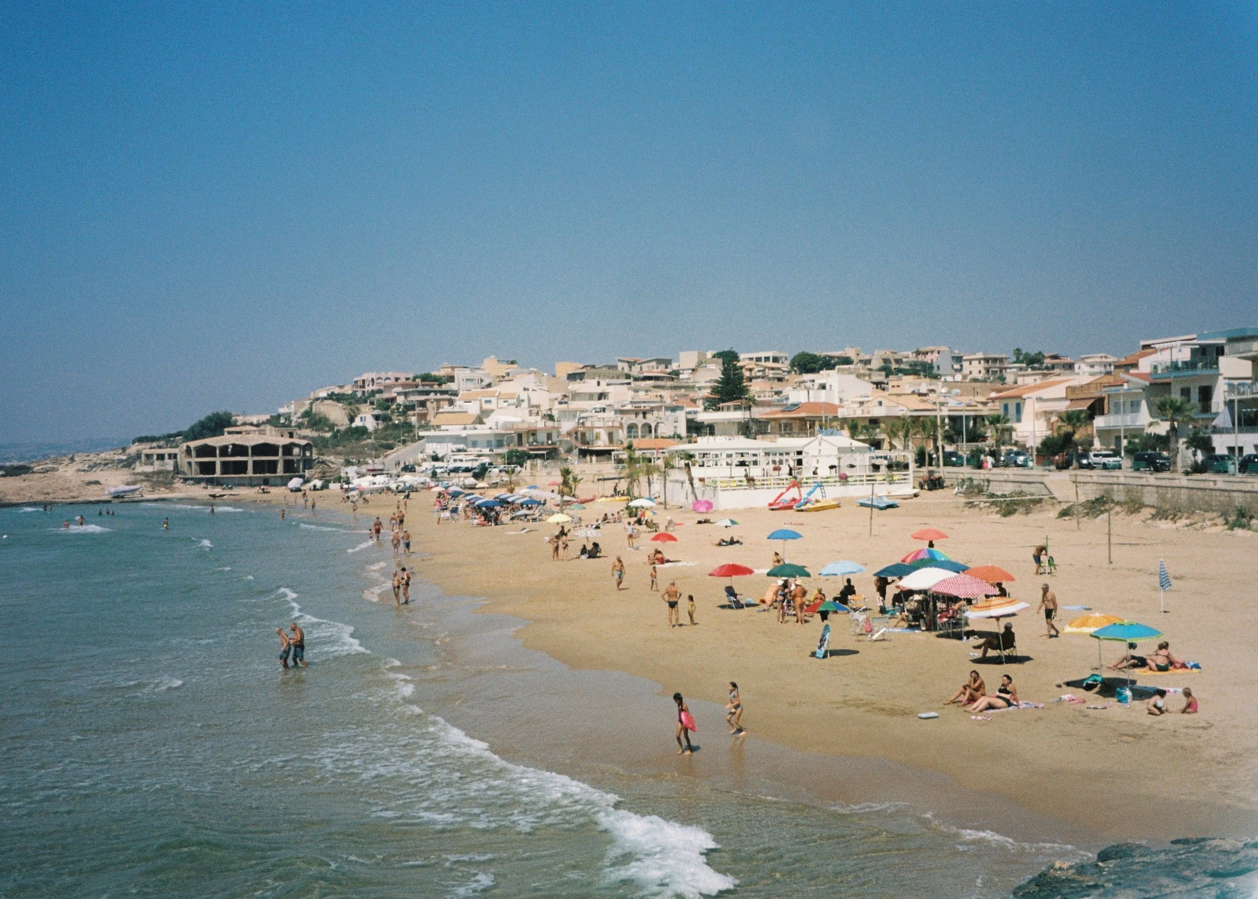 Beach scene with people relaxing under umbrellas, swimming, and walking along the shoreline, with a hillside of houses in the background.