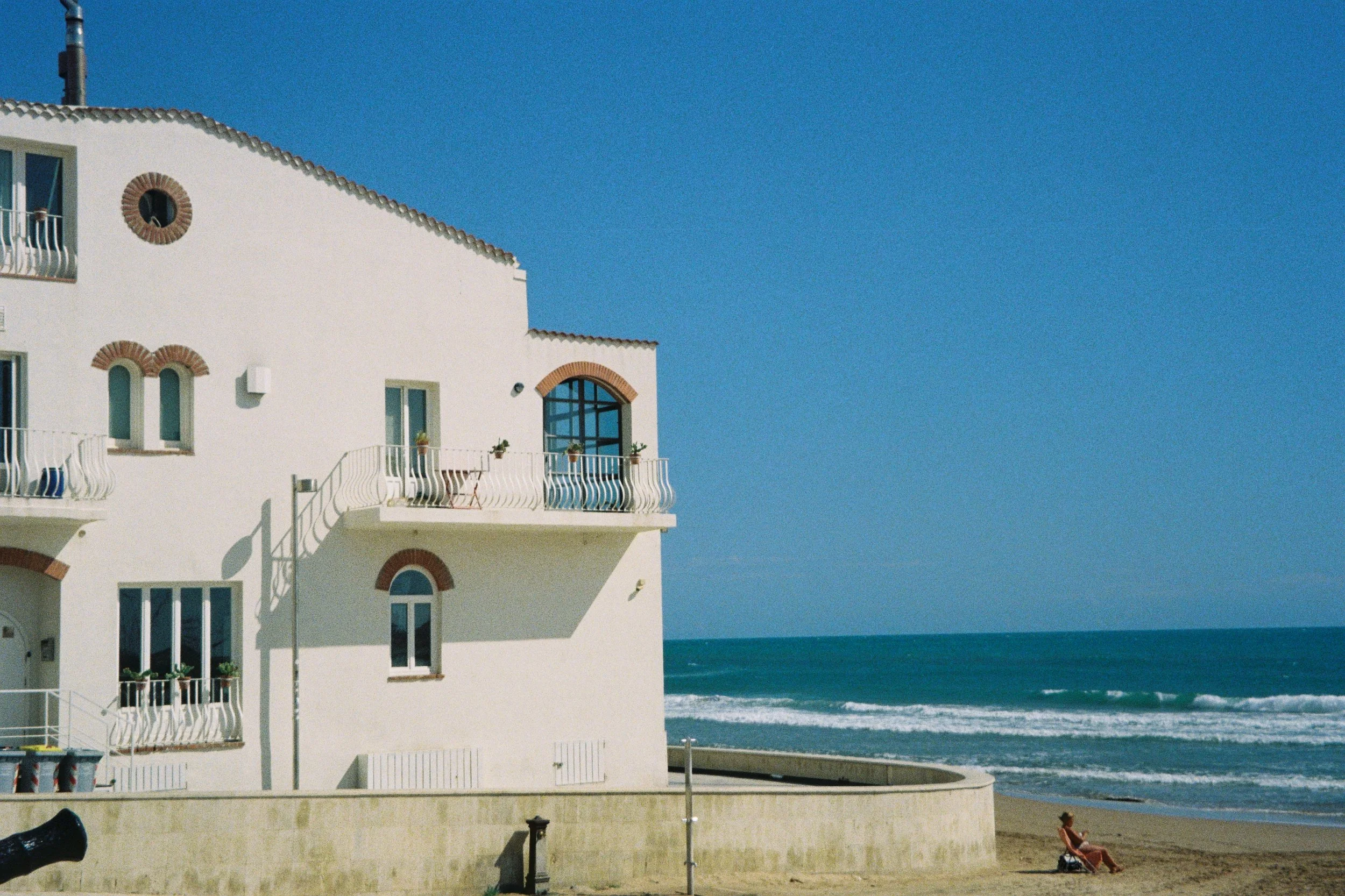 White multi-story beachside building with balconies, arched windows, and decorative brick accents under a clear blue sky, situated by the ocean.