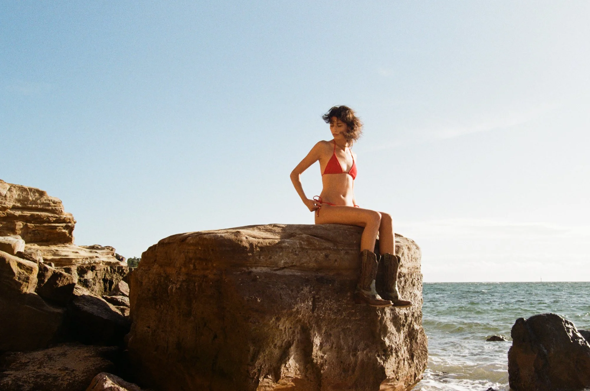 A woman sitting on a rocky shoreline in a red bikini, facing sideways with curly hair, at a beach under a clear sky.