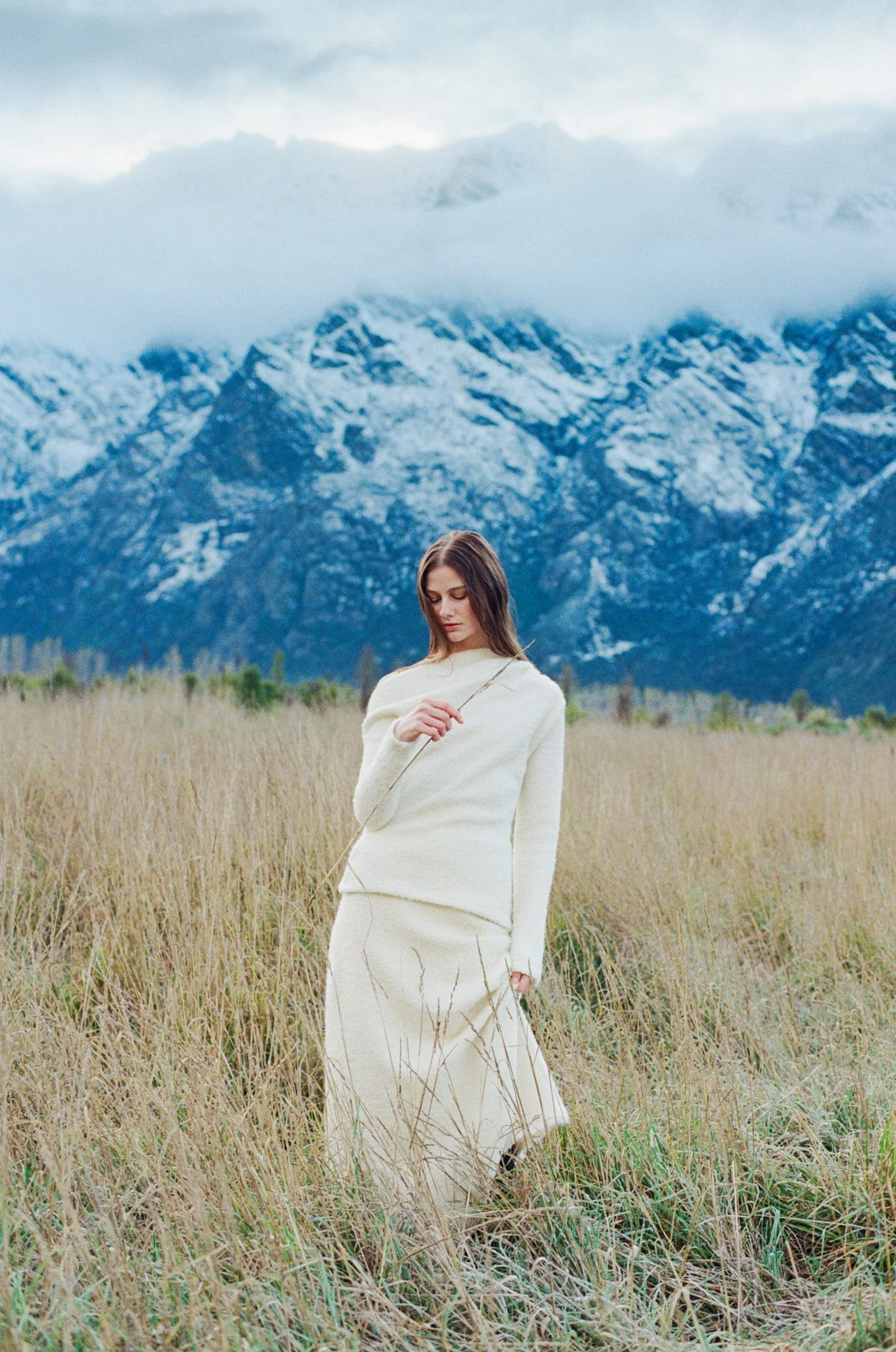A woman dressed in a cream-colored outfit stands in a field of tall grass, with snow-capped mountains and cloudy sky in the background.