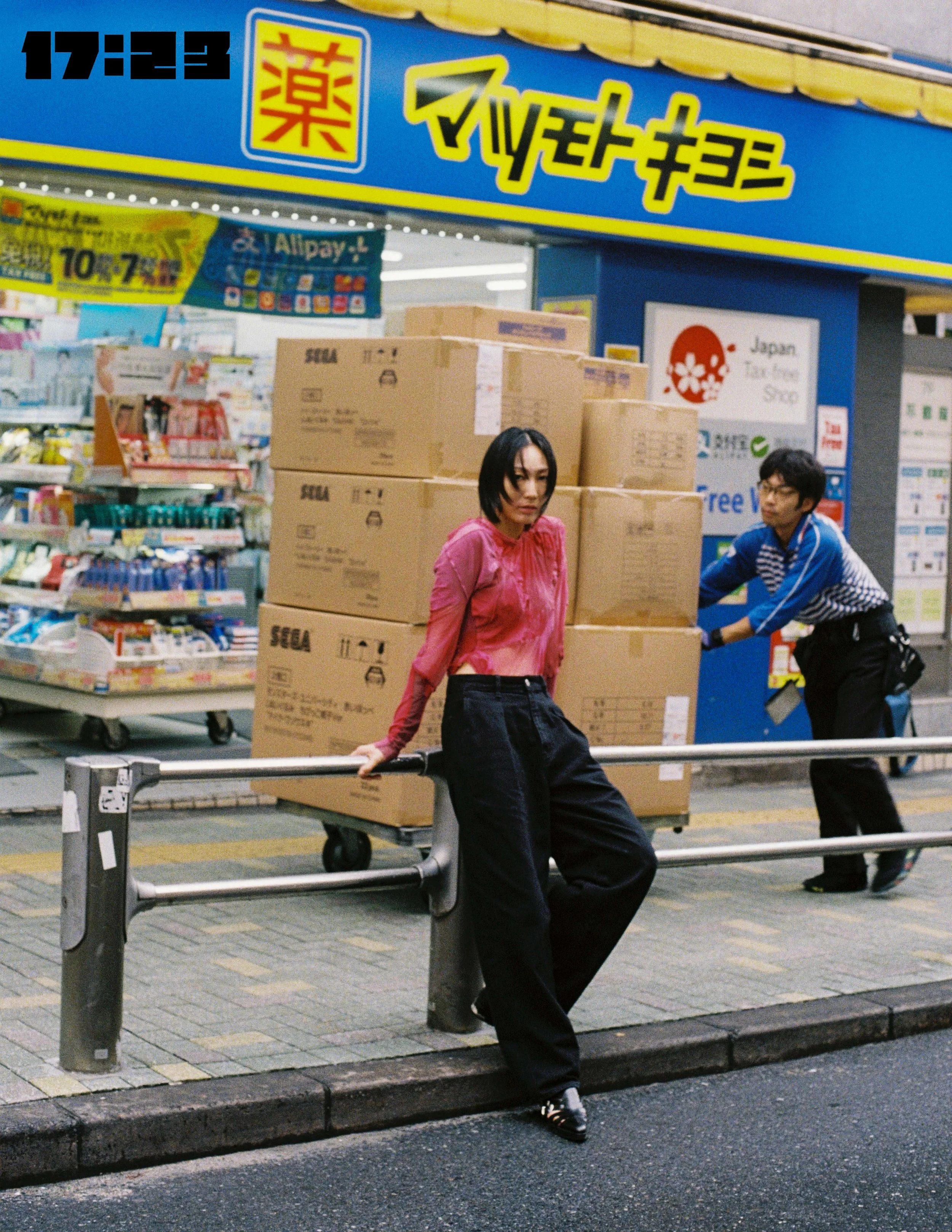 A young woman with short black hair and a pink shirt leaning on a shopping cart filled with boxes, standing on a city sidewalk with a woman in glasses and a striped shirt in the background, under a blue storefront sign with Japanese characters.