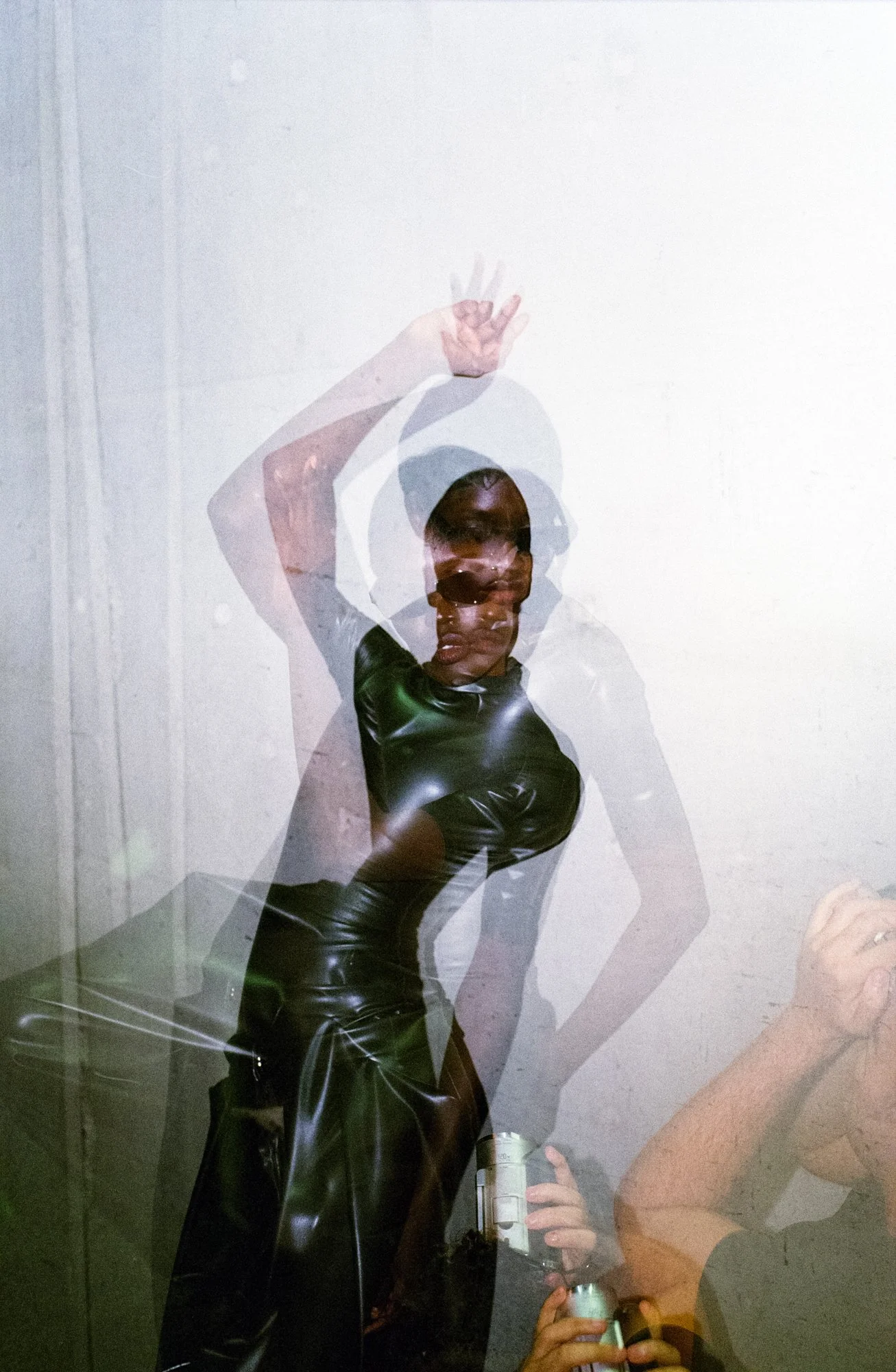 Multiple overlapping reflections of a woman with short hair and dark makeup, wearing a black shiny outfit, posing with one hand raised against a plain wall.