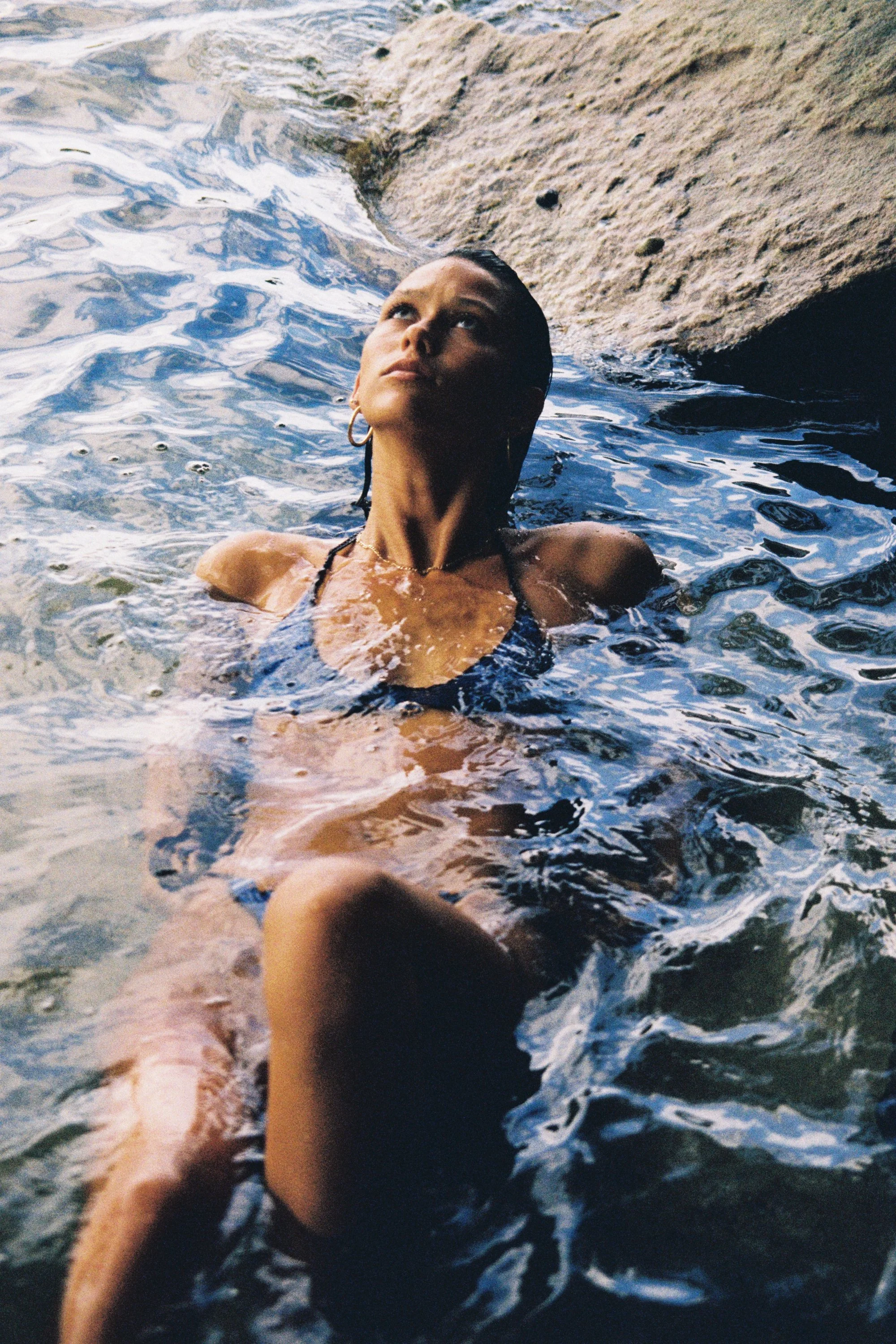 A woman with dark hair and hoop earrings in water, looking up, near a rock formation.