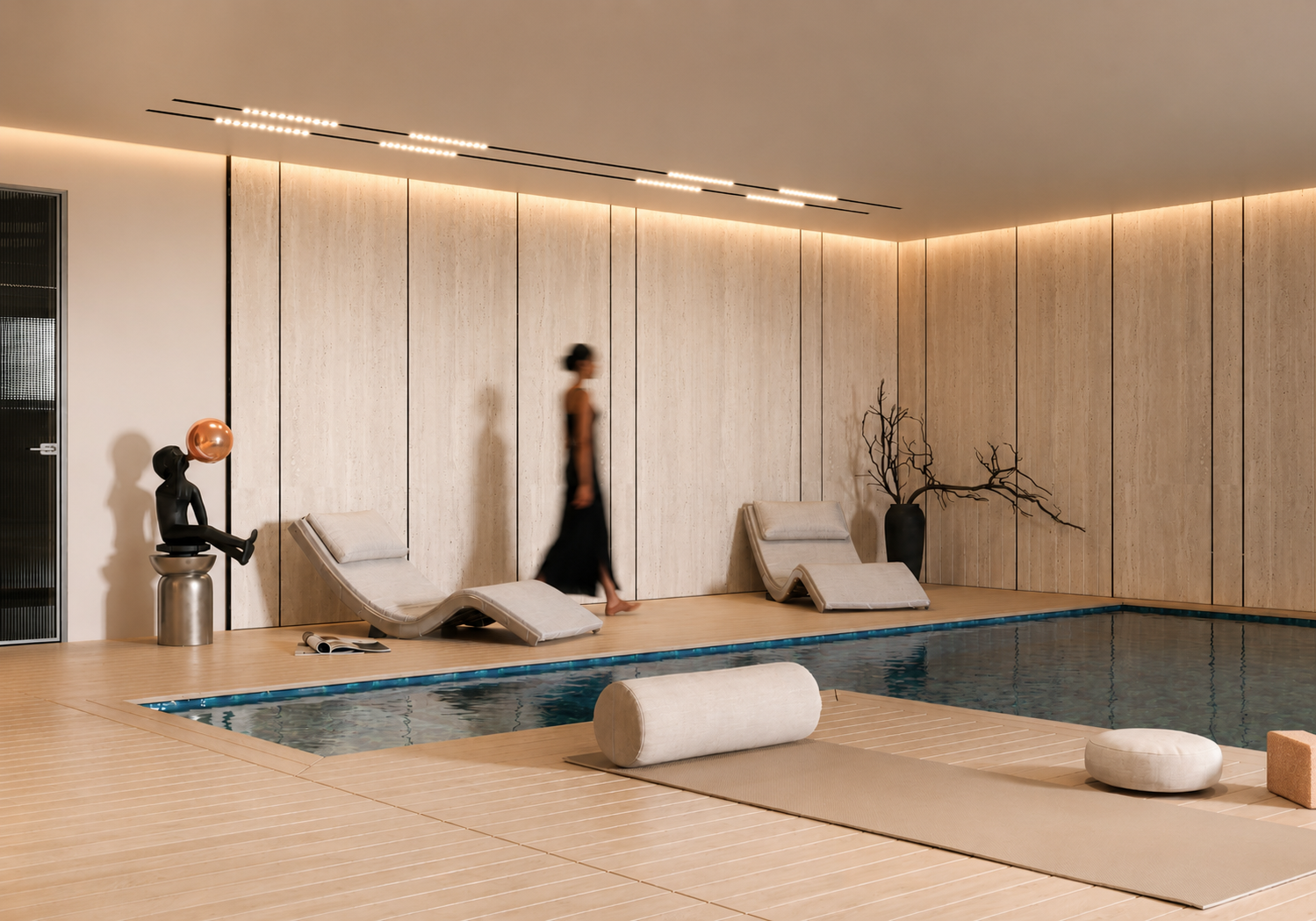 Modern indoor swimming pool area with beige lounge chairs, a decorative black sculpture with a copper sphere, a tall black vase with leafless branches, and a woman walking past. The walls are light-colored with vertical panels, and the ceiling has LED strip lighting.