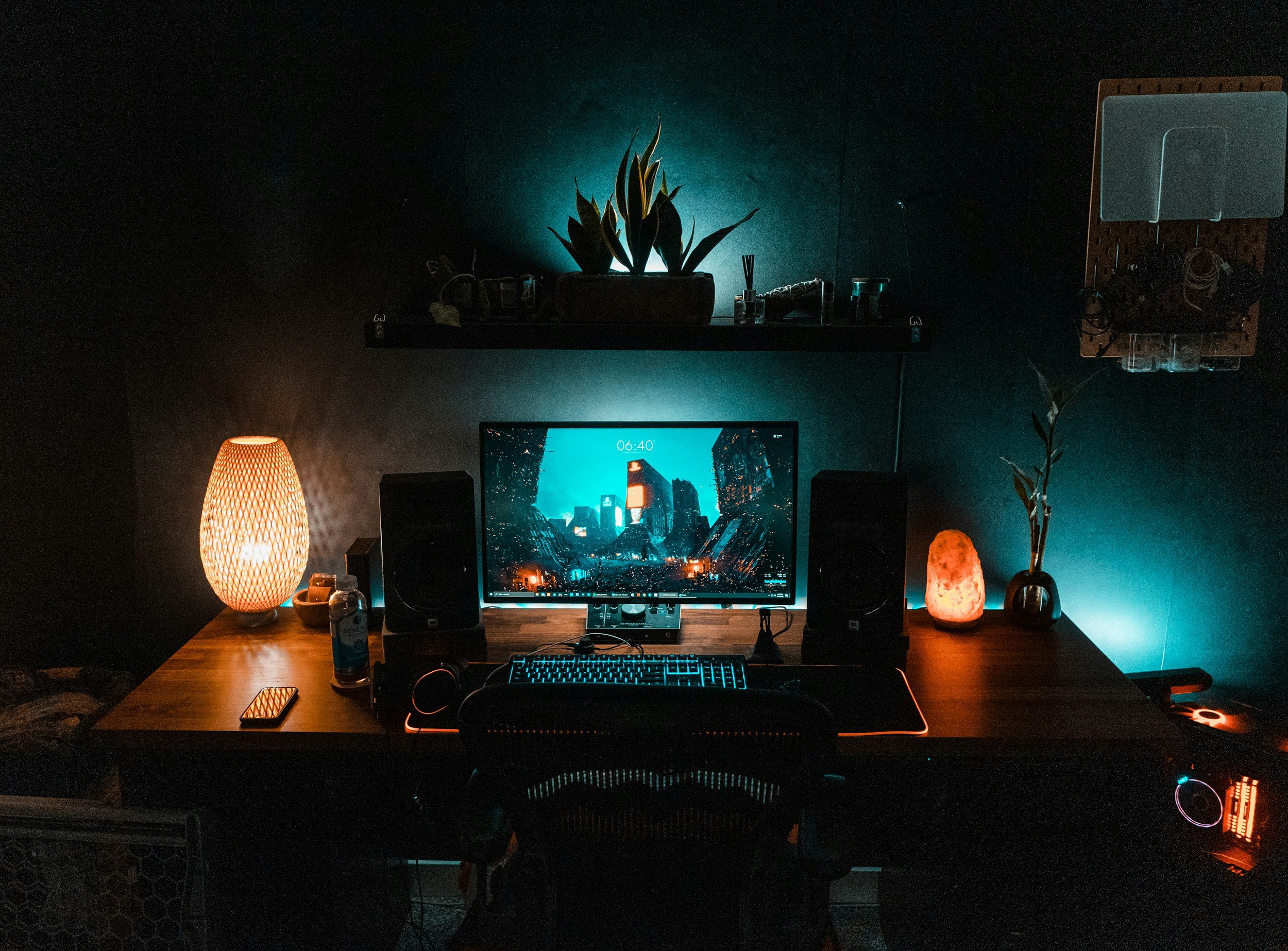 A neatly organized computer workspace with ambient lighting. The desk holds a monitor displaying a cityscape at night, a gaming keyboard, a mouse, and a smartphone. Warm-toned lamps on either side create a cozy atmosphere. A shelf above the desk contains plants, decorative items, and a diffuser, with a pegboard on the right for hanging accessories.