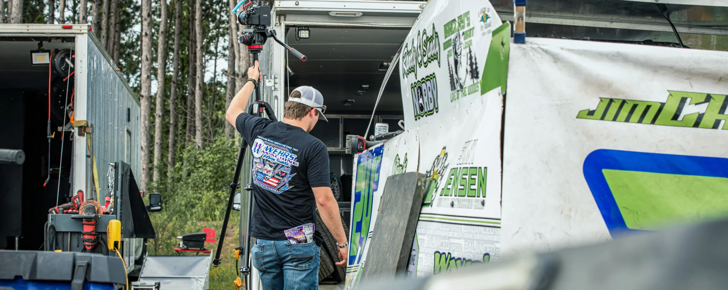 Person filming a race car on a tripod near a trailer in an outdoor setting.