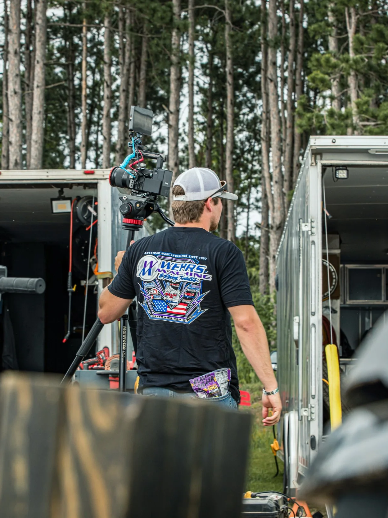 A man operating a film camera on a tripod outdoors, with trees in the background. He is wearing a black T-shirt with colorful racing graphics on the back, a gray baseball cap, and sunglasses, surrounded by equipment and storage units.