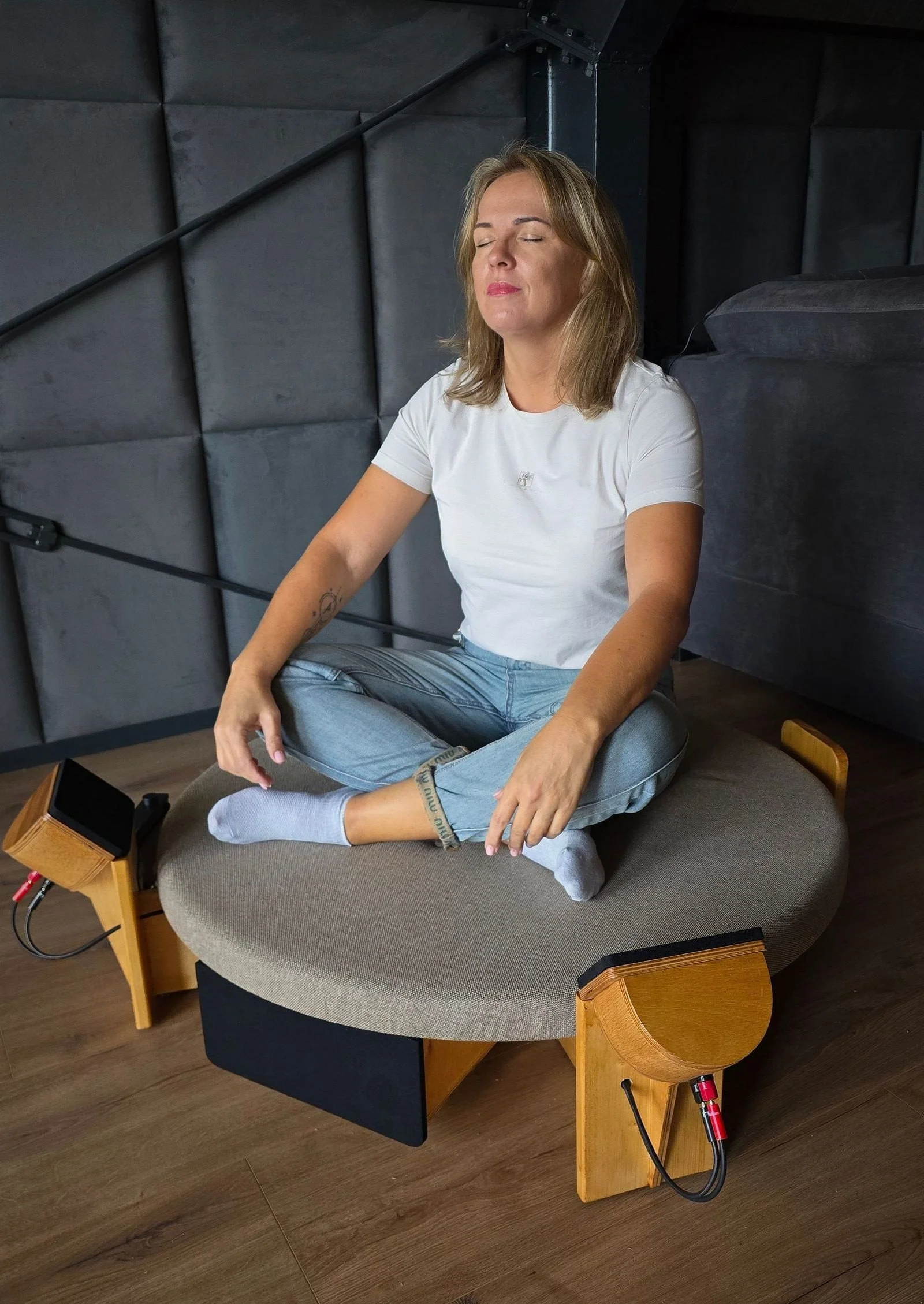 A woman practicing meditation in a seated cross-legged pose on a cushion, with eyes closed, in a room with dark walls and wooden flooring, connected to a nearby amplifier with cables.