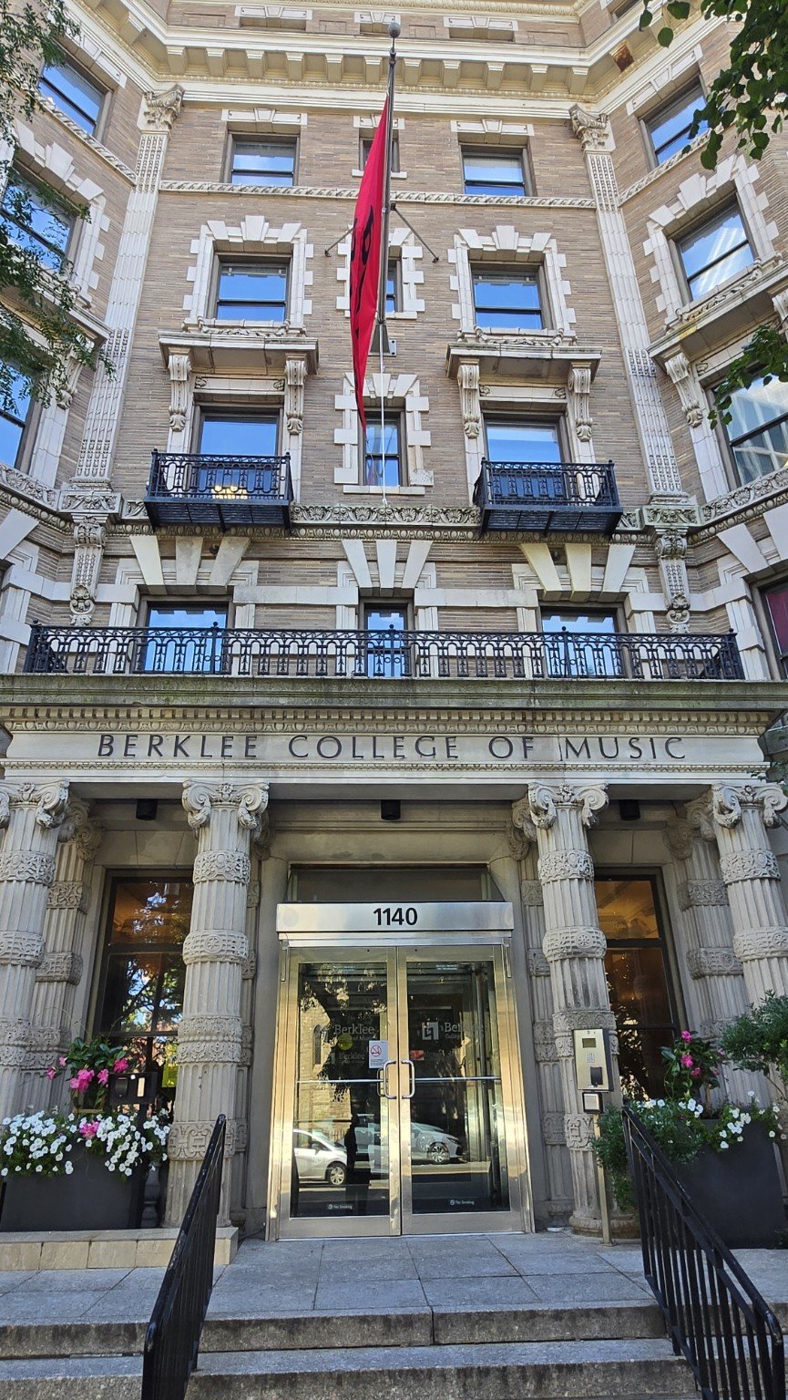 The building is Berklee College of Music, located at 1140, with a brick facade and ornate architectural details. There are balcony railings, a red vertical flag, and flowers in pots near the entrance.