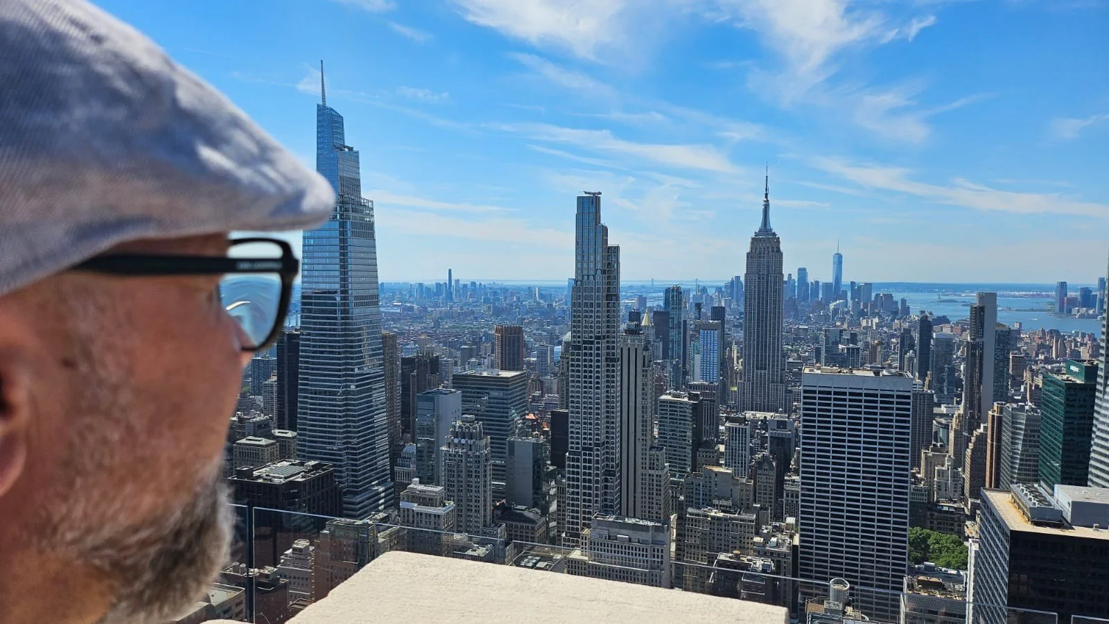 Close-up of a person's face in profile, wearing glasses and a grey hat, overlooking the New York City skyline with notable skyscrapers including the Empire State Building, on a clear day.