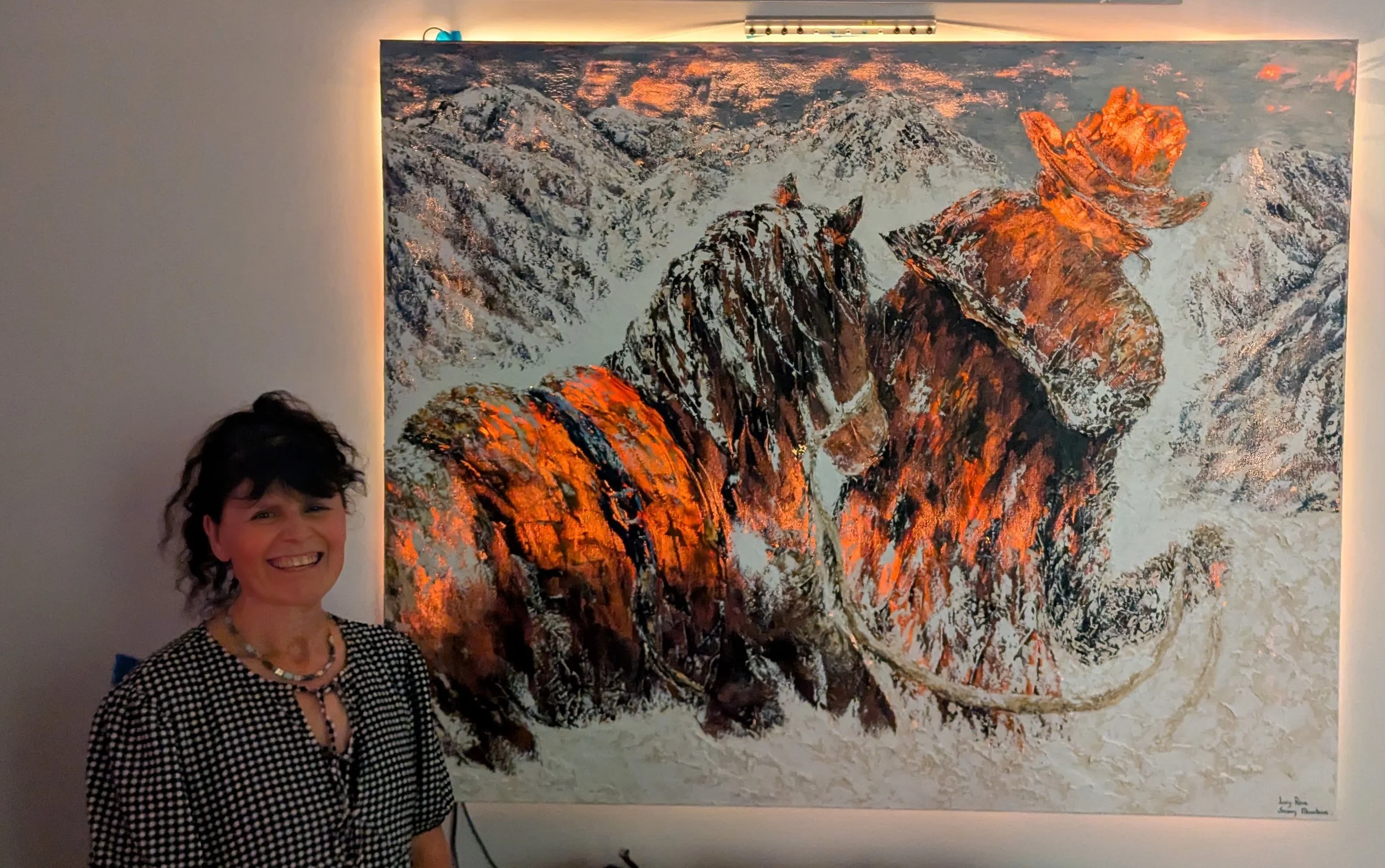 A woman with dark, curly hair smiling at an art exhibit, standing in front of a large abstract painting of a mountain landscape with warm tones and textured brushstrokes.
