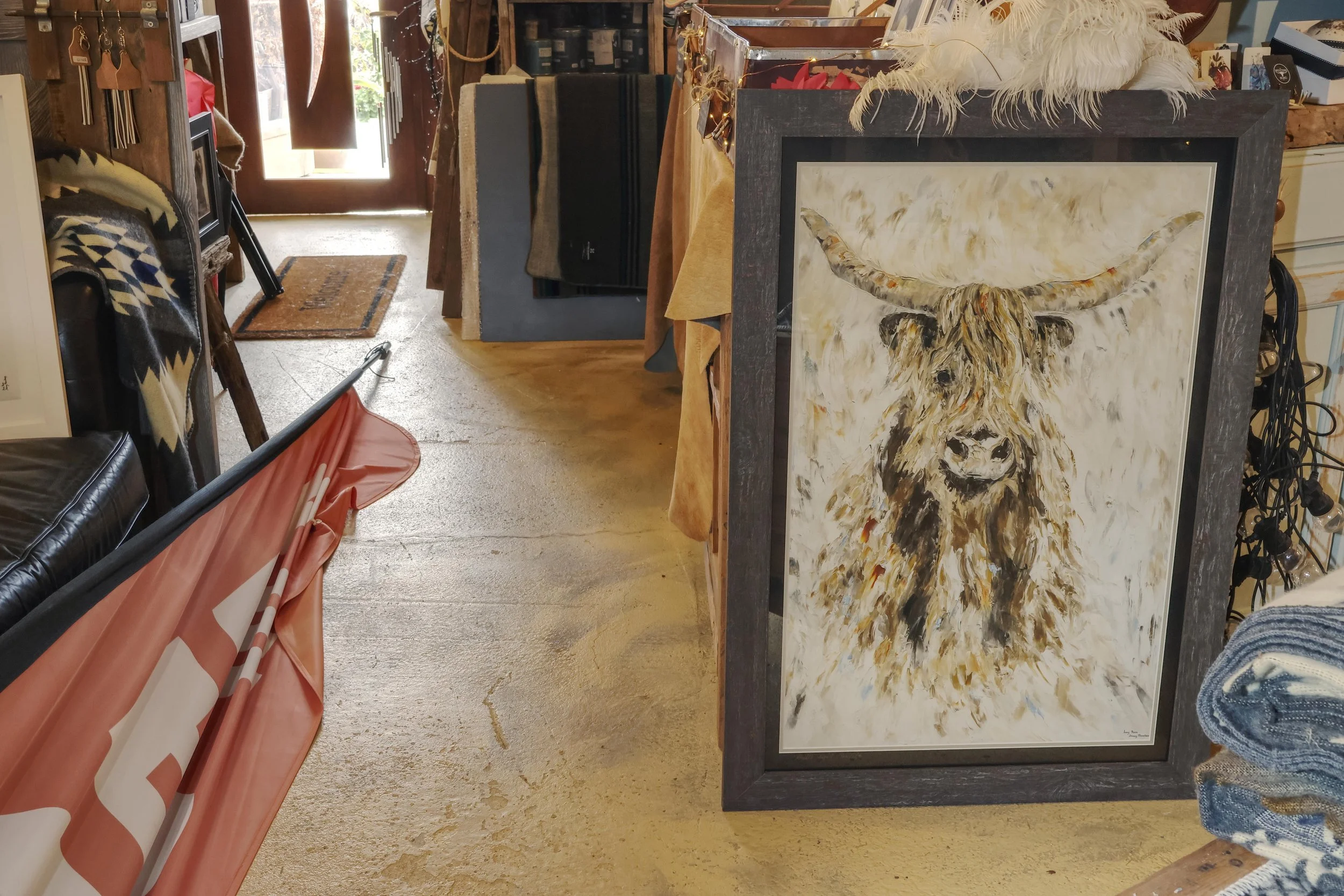 A framed painting of a bovine head, with a feathered headdress, leaning forward. The painting has a light background and is displayed upright among other items in an indoor setting.