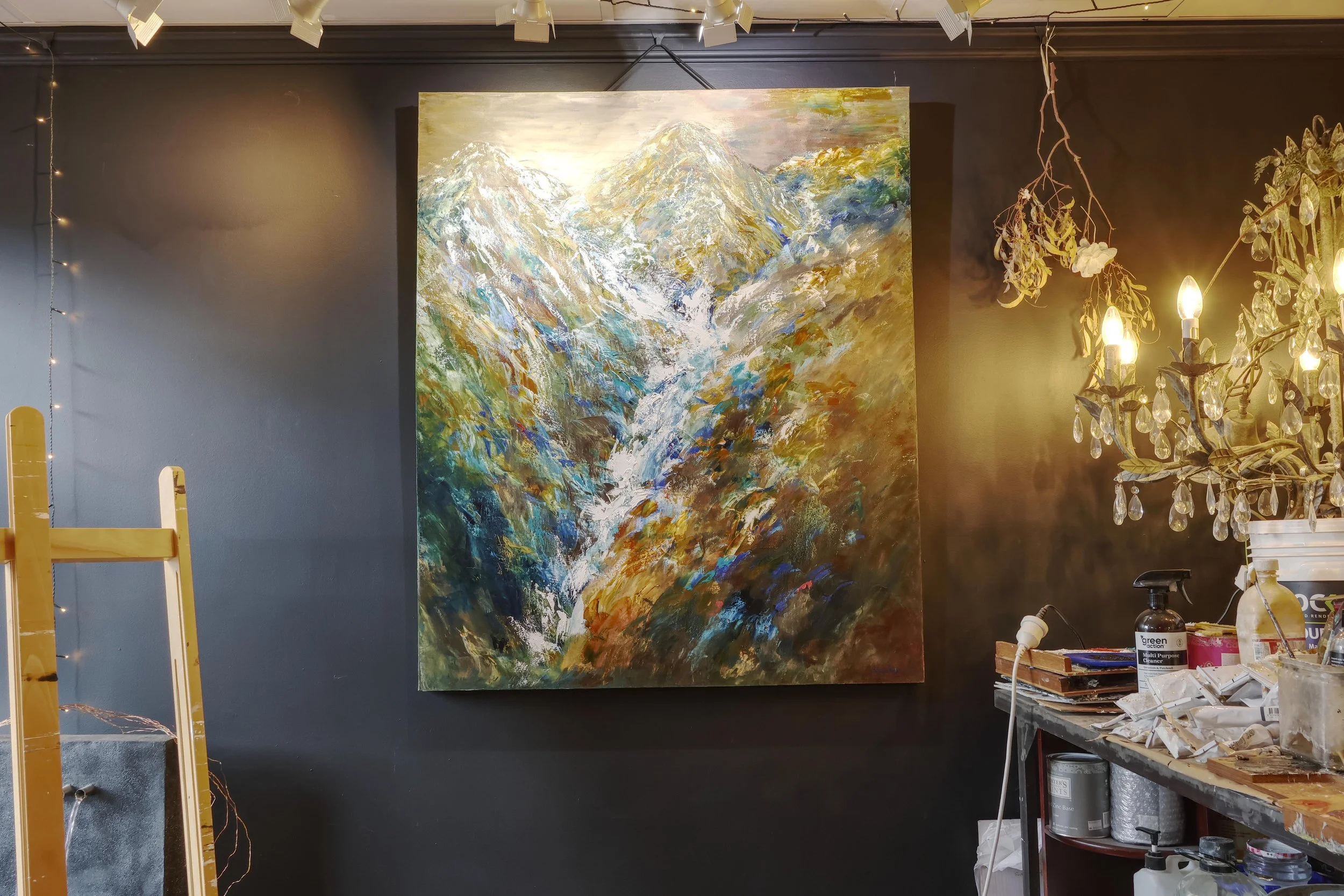 An abstract painting of a mountain landscape on canvas, displayed on a dark wall in an art studio. The scene features rugged mountains with snow-capped peaks, painted with vibrant and textured brushstrokes in blues, greens, whites, and earth tones. T