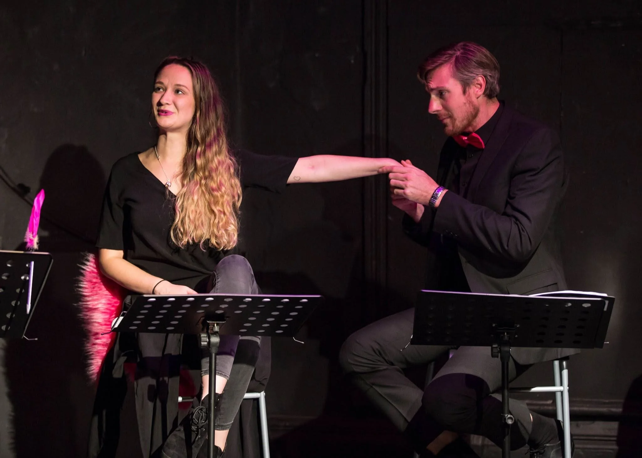 A man in a black suit with a red bow tie is touching the arm of a woman with long, wavy hair who is sitting on a chair. The woman is wearing a black top and grey pants, and is smiling. There are music stands and chairs in front of them, indicating a 