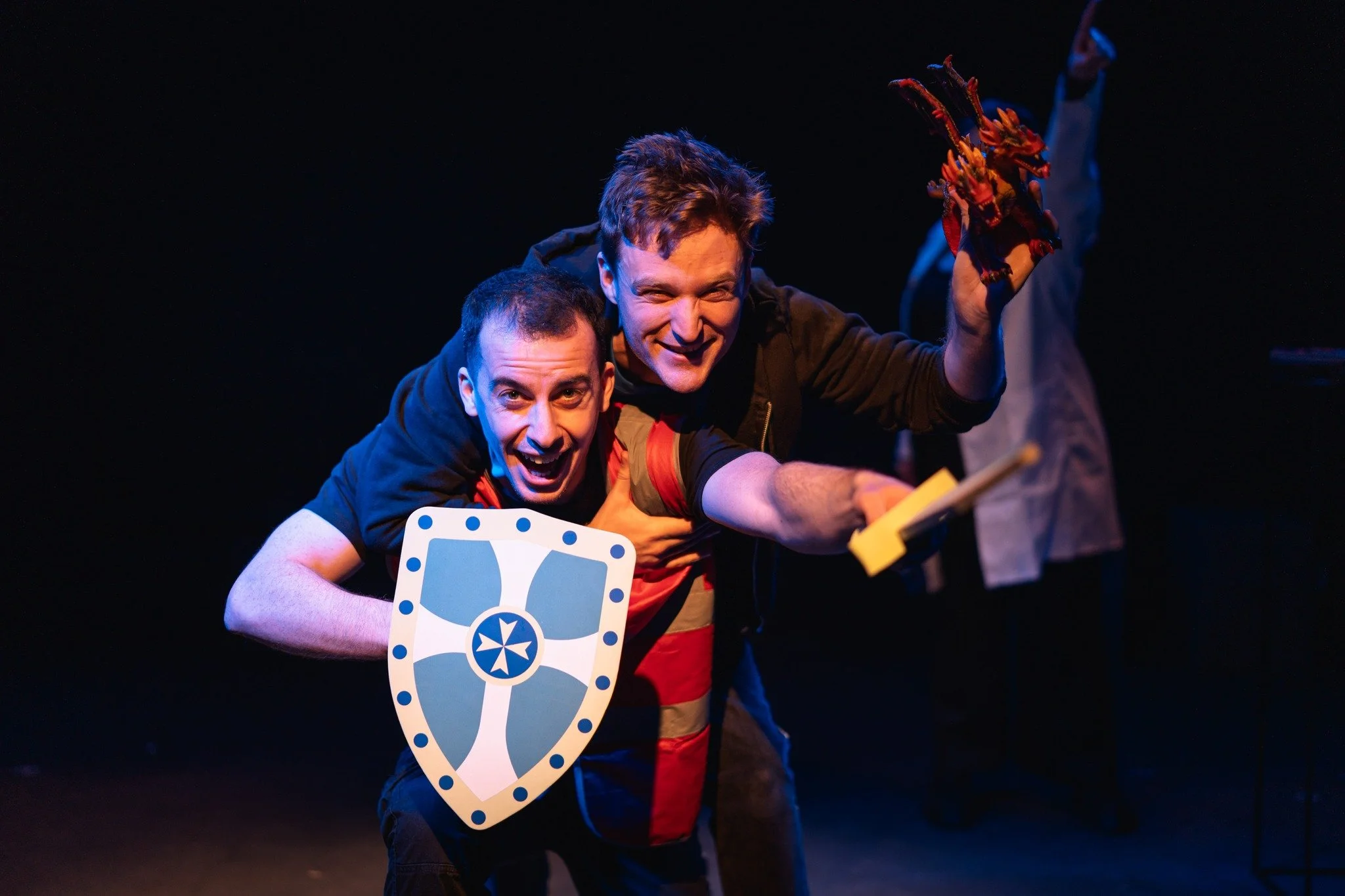 Two men on stage participating in a performance or game. One man is holding a shield and a sword and they both look excited and smiling. The scene is dark with stage lighting highlighting the men.