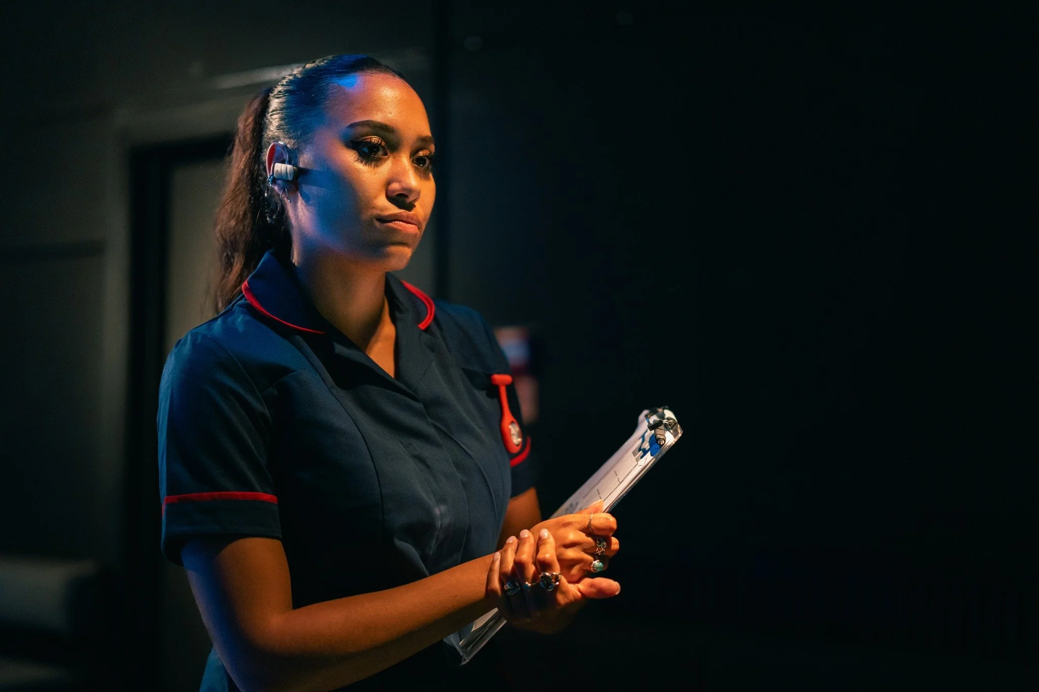 A woman in a dark blue uniform with red accents stands in a dimly lit room, holding a clipboard and a pen, with an earpiece in her right ear, appearing focused.