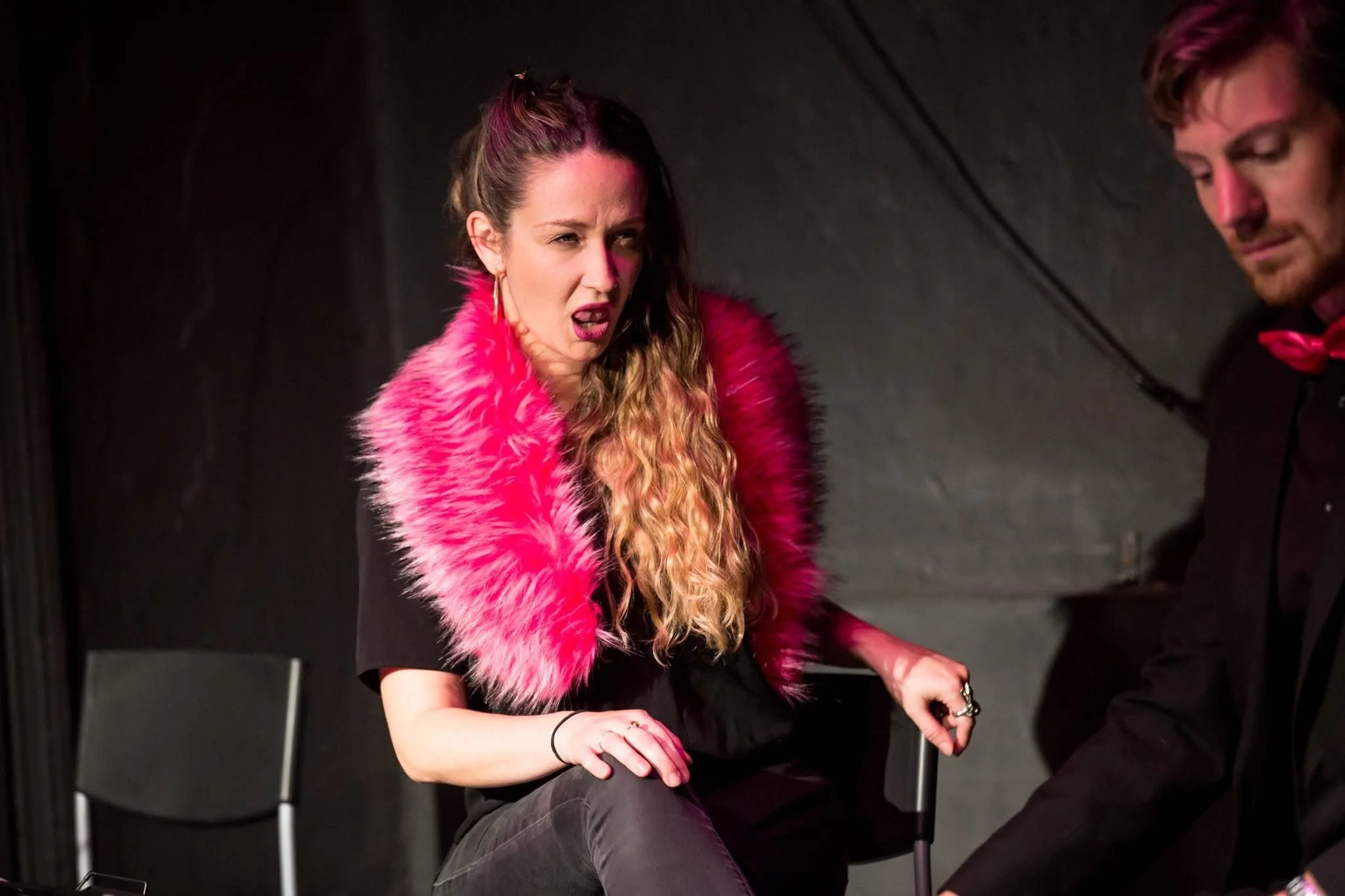 A woman with long, curly hair wearing a bright pink faux fur coat, sitting and making a confused or annoyed facial expression, with another person partially visible next to her.