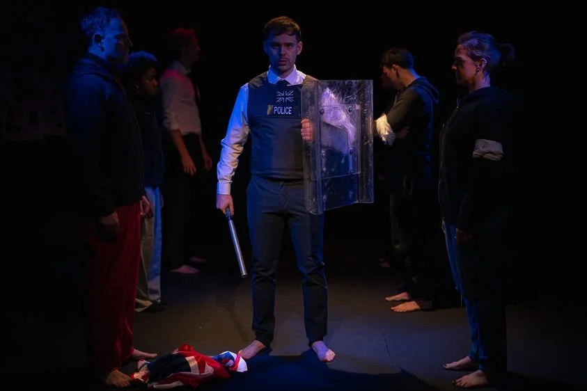 A group of people standing in a dark room, with one man in the center dressed as a police officer holding a shield and baton, surrounded by others who are barefoot and facing him.