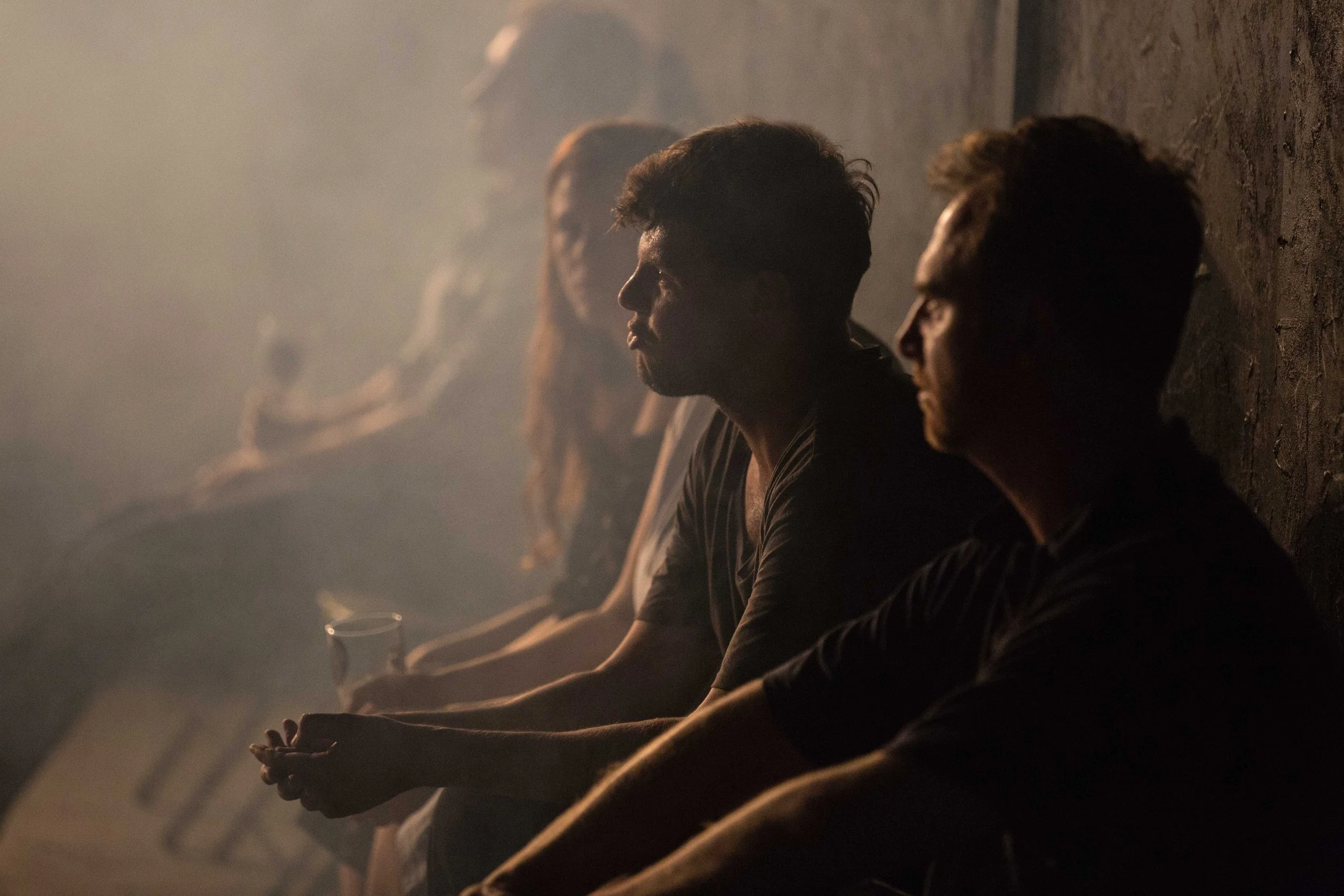 Four men sitting against a wall in a dimly lit space. The man closest to the camera has gloves on and is holding his hands together, looking straight ahead. The others are seated behind him, facing forward.