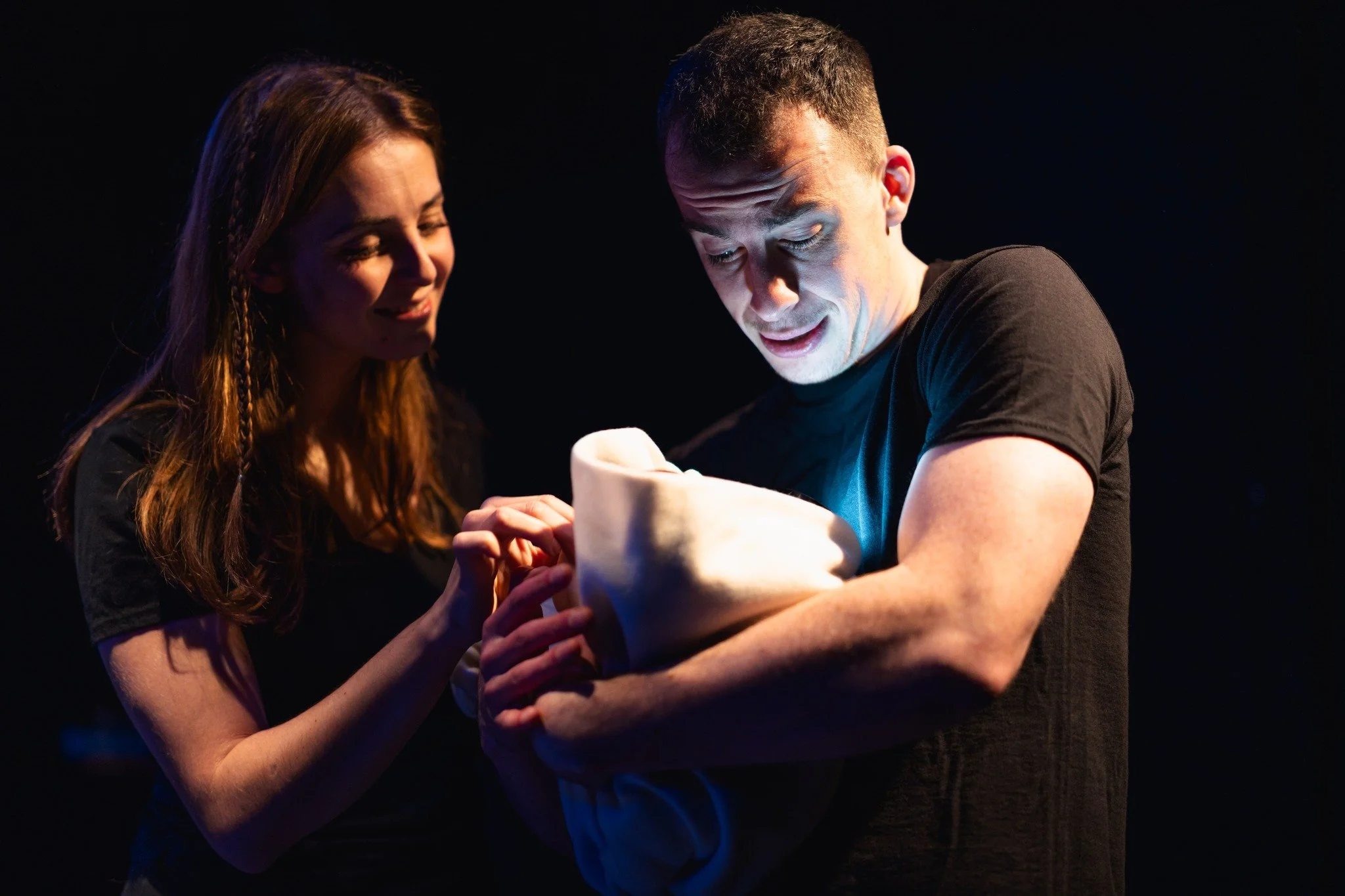 A young woman and a man are holding a baby together, smiling and looking at it in a dark setting with stage lighting.