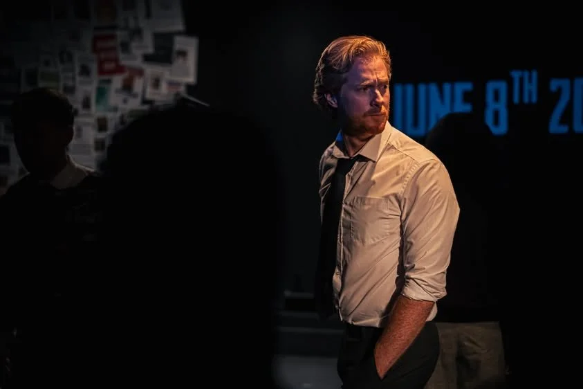 A man with red hair and a beard wearing a beige shirt and black tie, standing in a dark room with a blurred background, looking to his right. There is blue text on the wall behind him that reads 'June 8th 20...'.