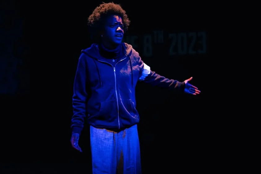 A person with curly hair standing on stage lit with blue light, gesturing with one arm extended.