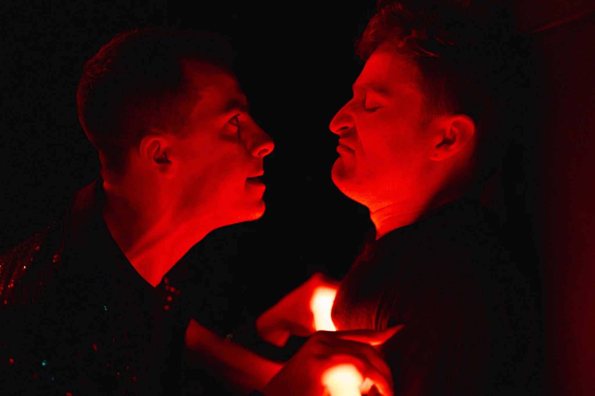 Two people face to face in a dark setting, combative,  illuminated by red lighting, almost touching foreheads, 