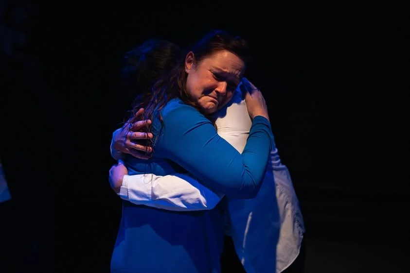 A womand and a young man hugging emotionally displayed on a dark stage, one woman visibly crying.