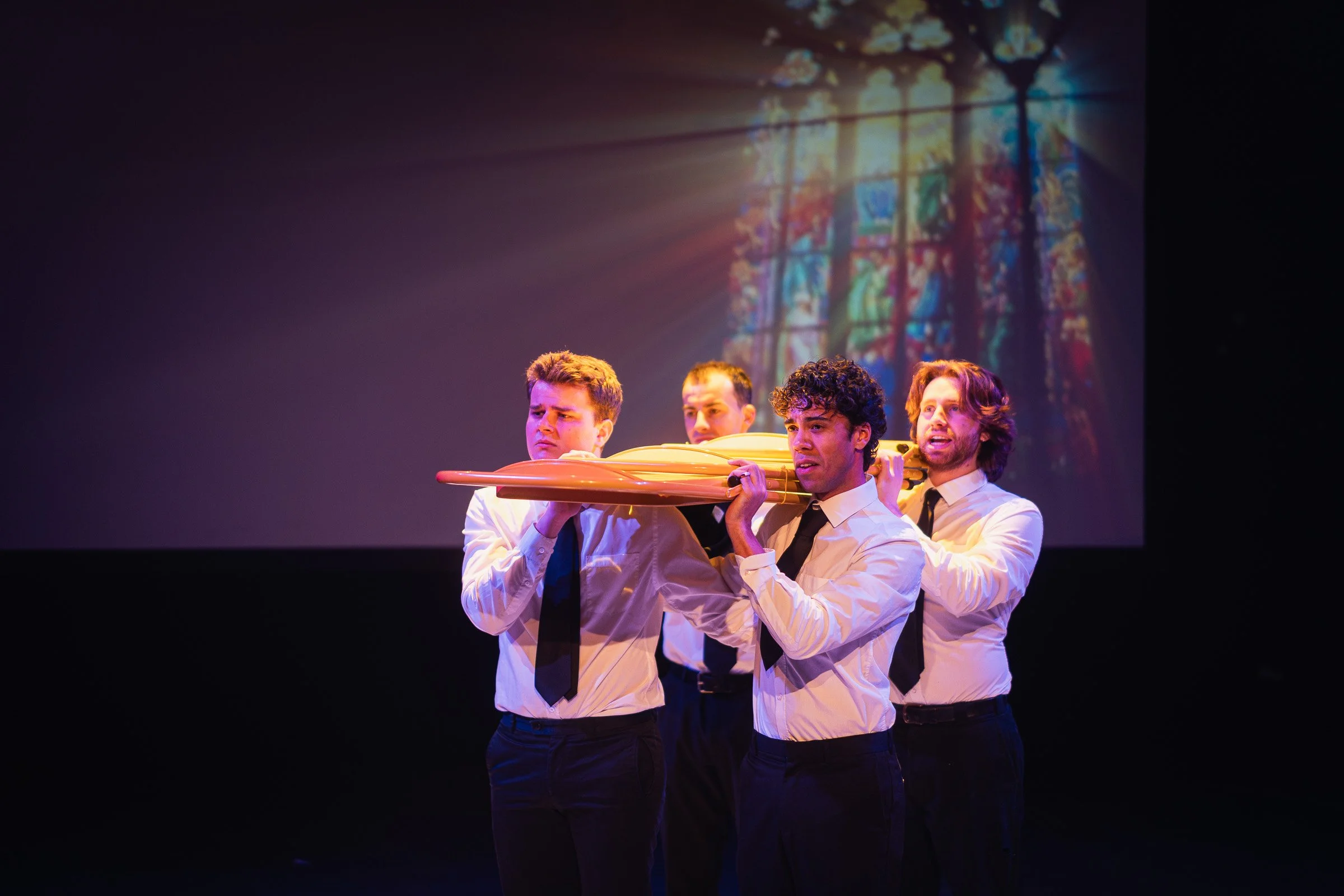 Four men dressed in white shirts and black ties carrying an impression of a coffin on stage, with a stained glass window projection in the background.