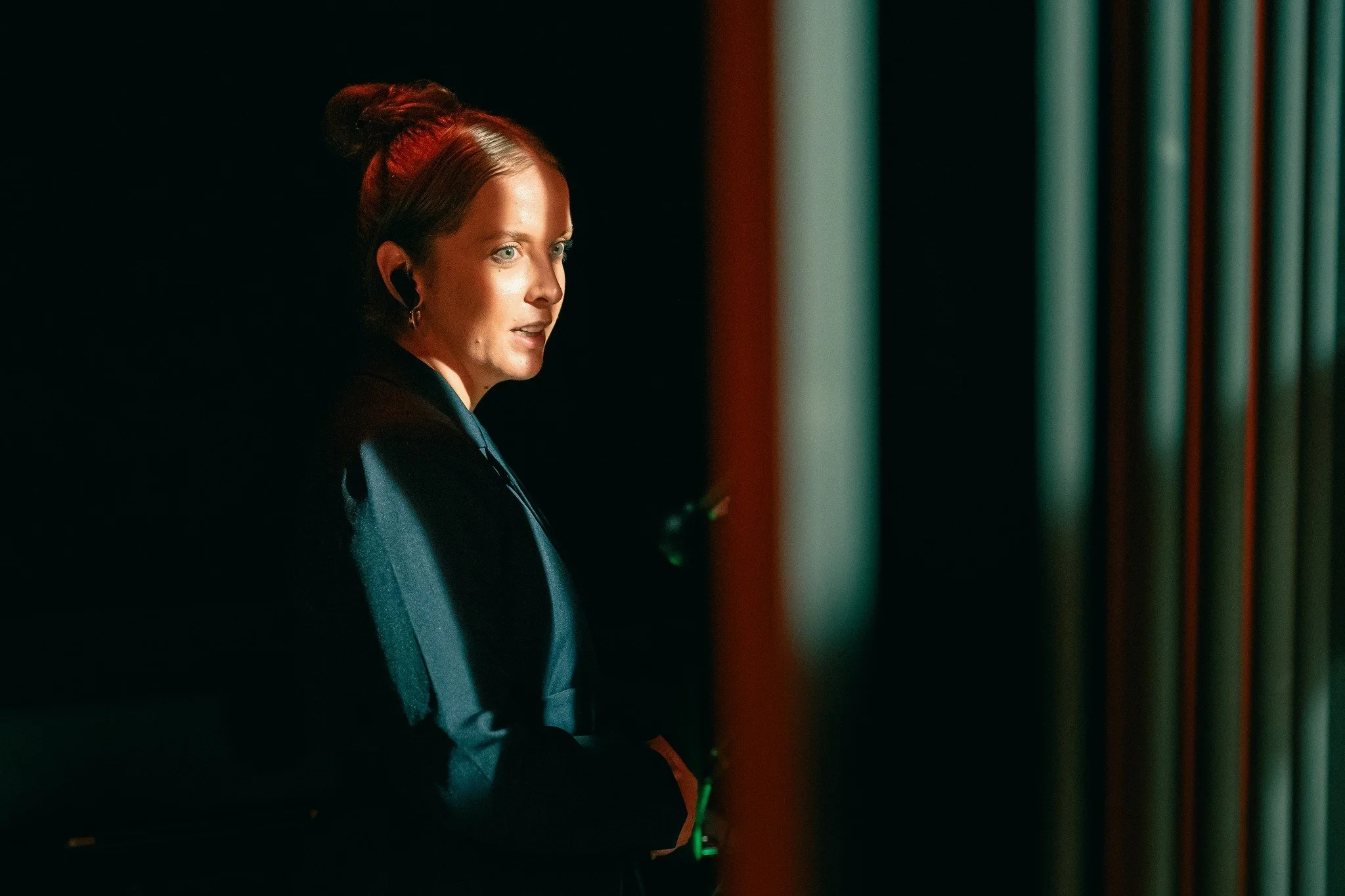 A woman with red hair styled in an updo, wearing earbuds and a dark suit, standing in a dimly lit room with vertical green lights in the background.