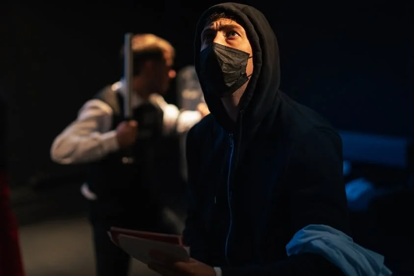 A person wearing a black hoodie and face mask, holding papers, in a dark setting with another person blurred in the background.