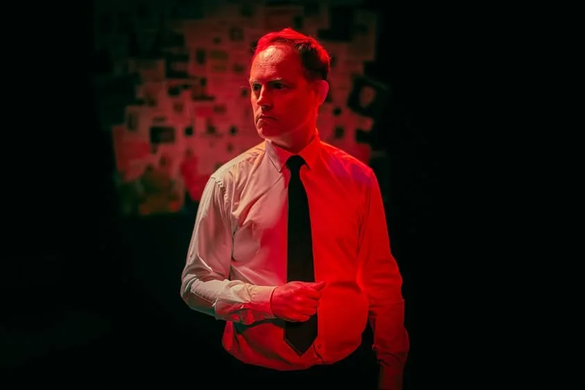 A man in a white dress shirt and black tie stands in a dimly lit environment with intense red lighting, with a blurry wall in the background.