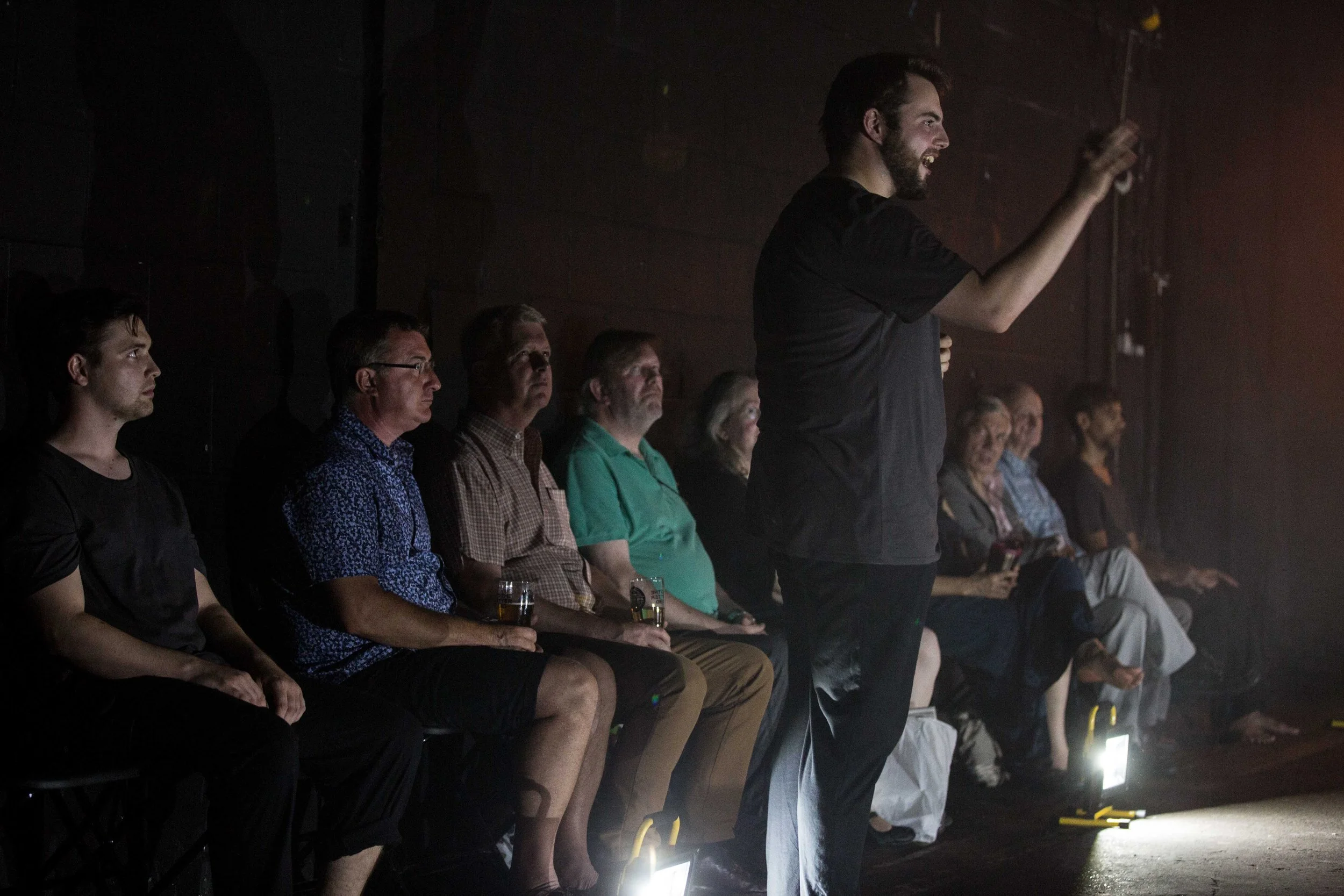 A group of people seated in a dark room with a man standing and speaking, illuminated by a light at the front; some attendees are holding glasses of beer.